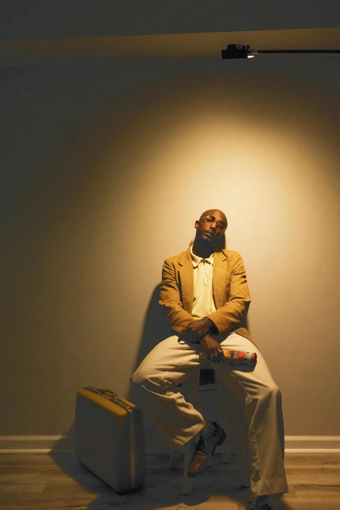 A man with glasses sitting on a white chair against a plain wall, holding a colorful object, wearing a beige blazer, white shirt, and khaki pants, with a yellow briefcase on the floor nearby.