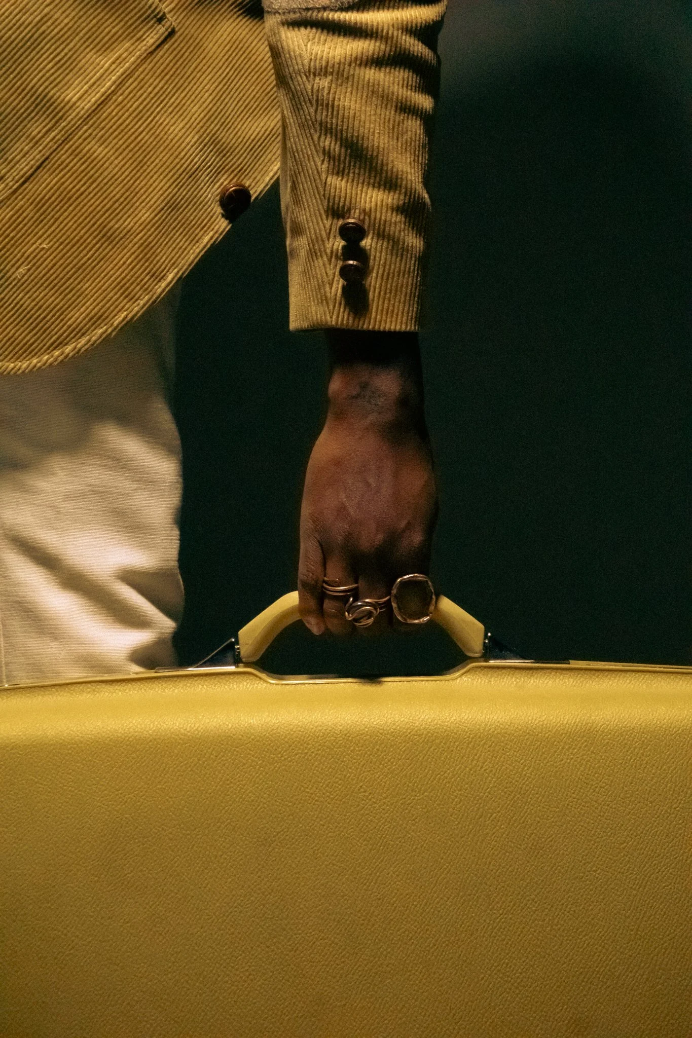 Close-up of a person holding a yellow suitcase, wearing a beige corduroy jacket with black buttons and rings on their fingers.