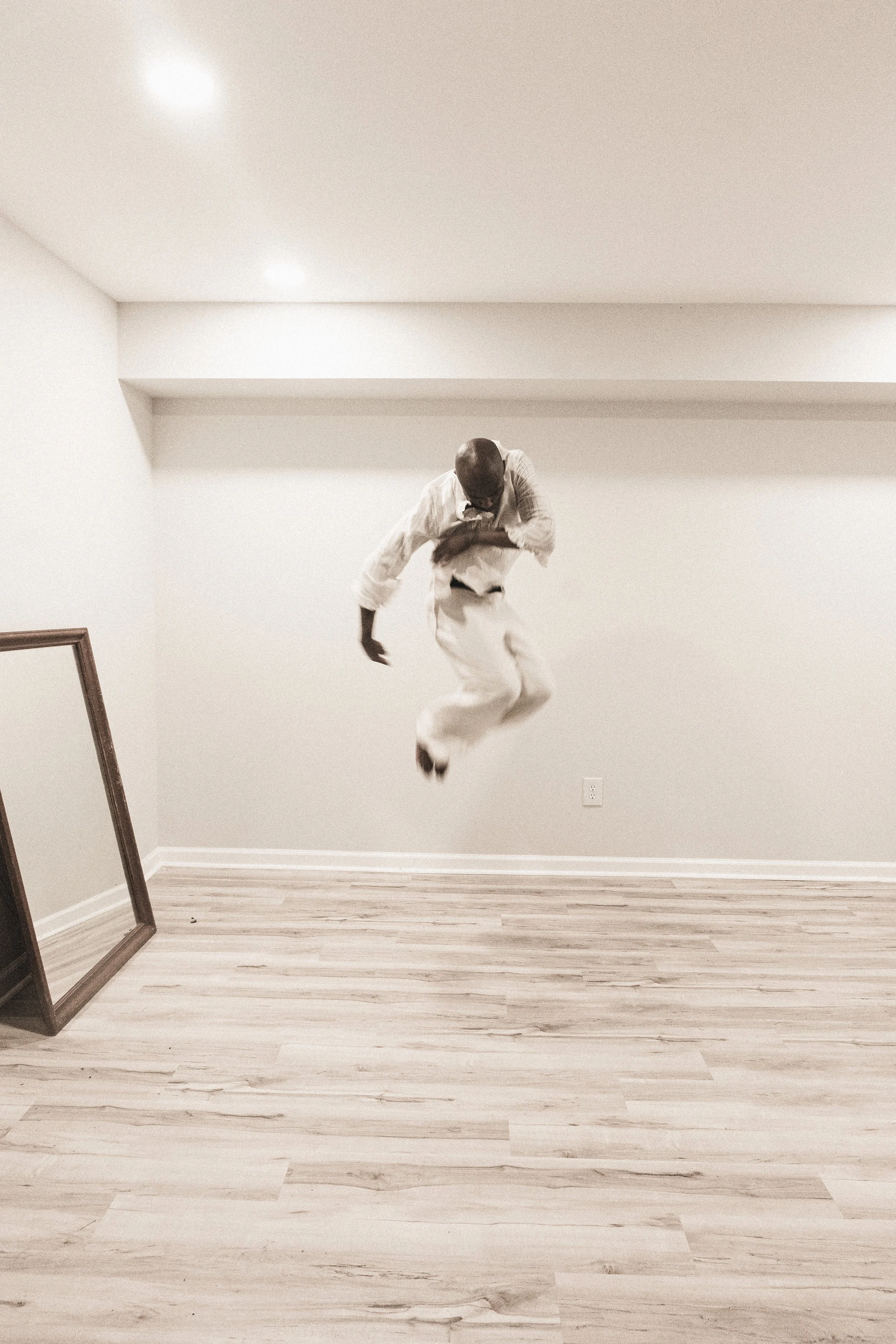 A person is mid-air, jumping in a room with beige walls and wooden flooring, with a large mirror leaning against the left wall and ceiling lights overhead.