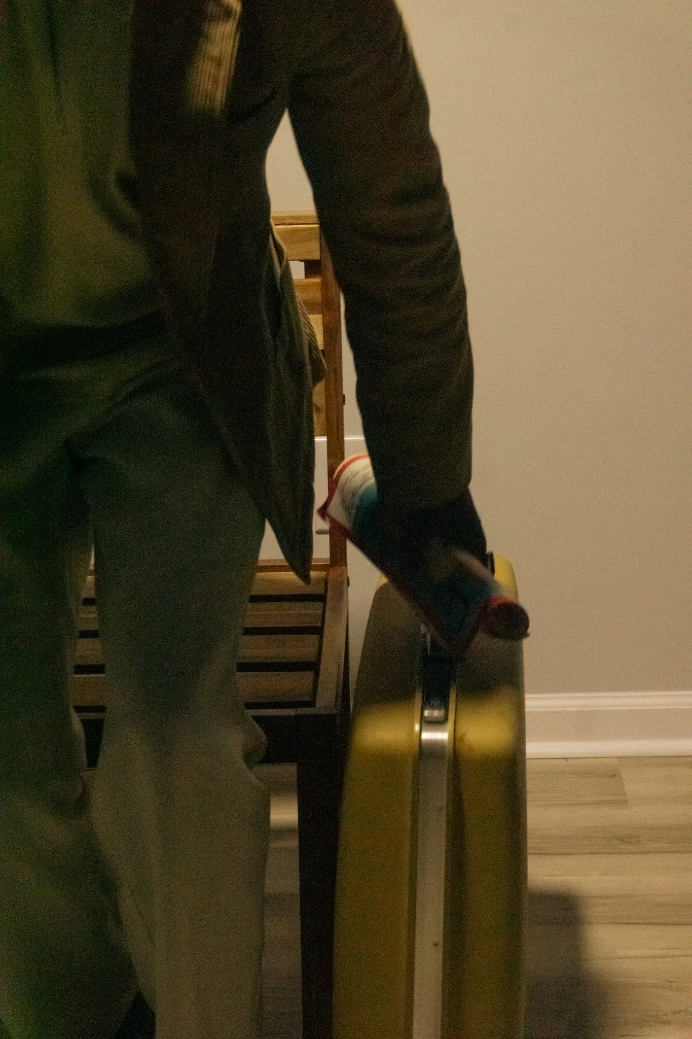 A reflection of a person taking a mirror selfie in a room with beige walls and wooden flooring. The person is wearing dark clothing and is holding a shoe in front of them.