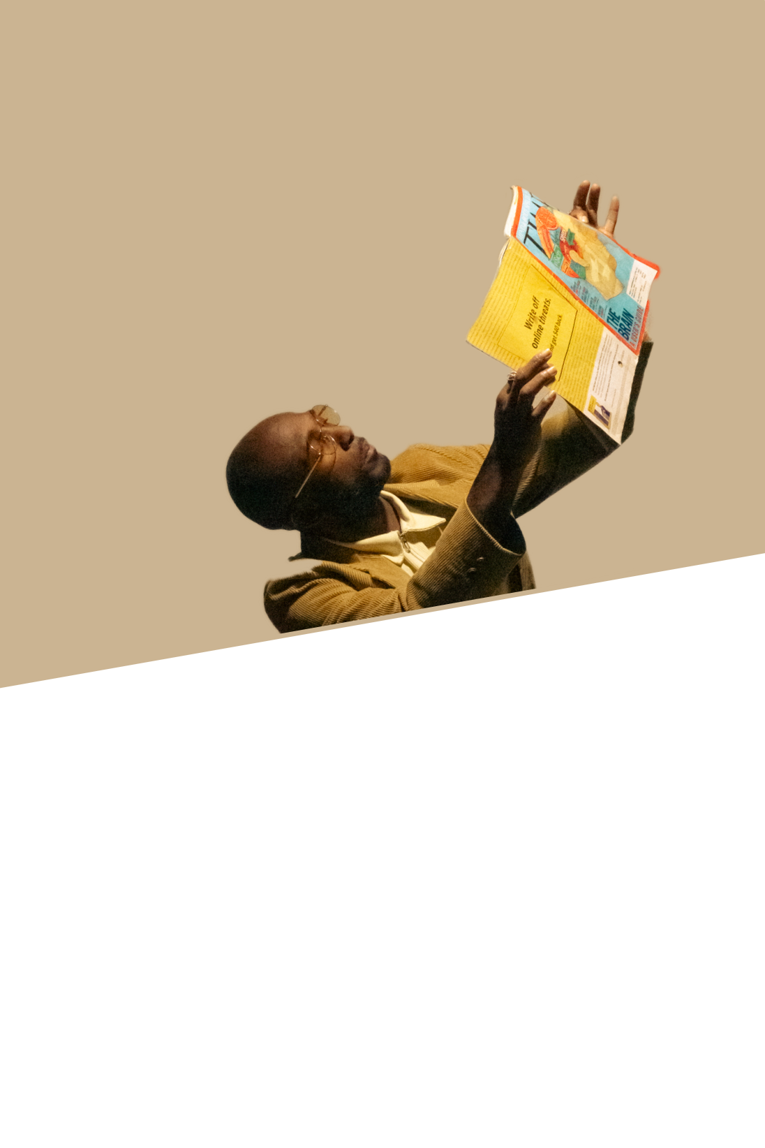 A man wearing glasses and a brown suit reading a magazine or newspaper against a plain background.