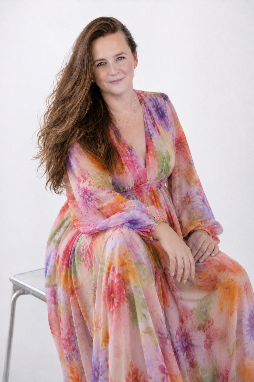 A woman with long, wavy brown hair wearing a colorful, floral, long-sleeved dress sitting on a silver metal chair against a plain white background.