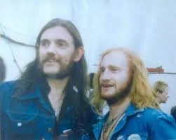 Two men with long hair and beards standing outdoors, wearing denim jackets, smiling.