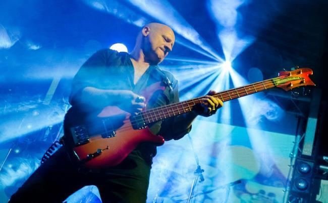 Musician playing an electric bass guitar on stage with bright blue lighting and a starburst in the background.