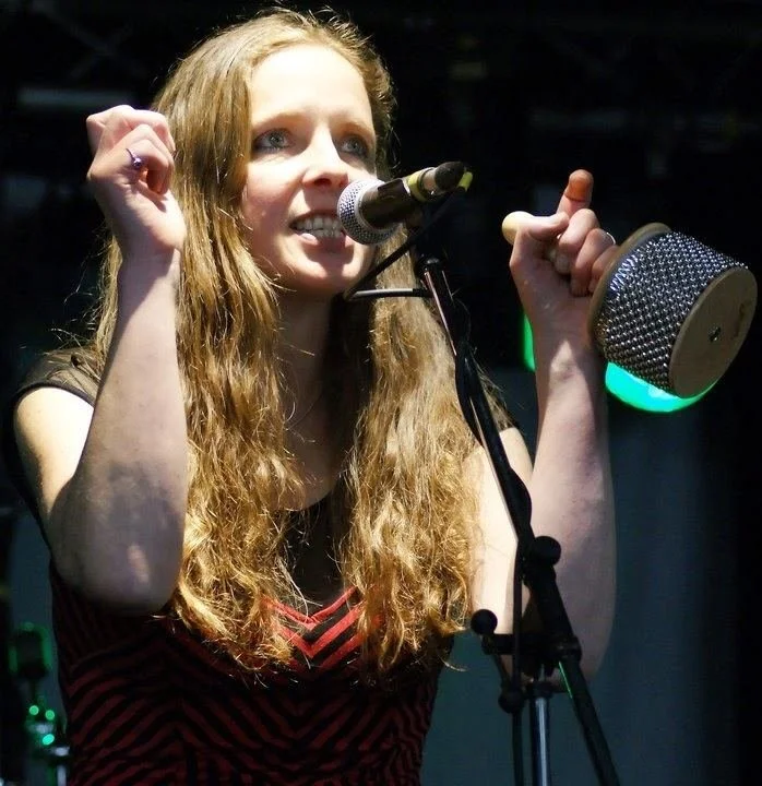 A woman with long, curly blonde hair singing into a microphone on stage, holding a drumstick in her right hand, with green stage lights in the background.