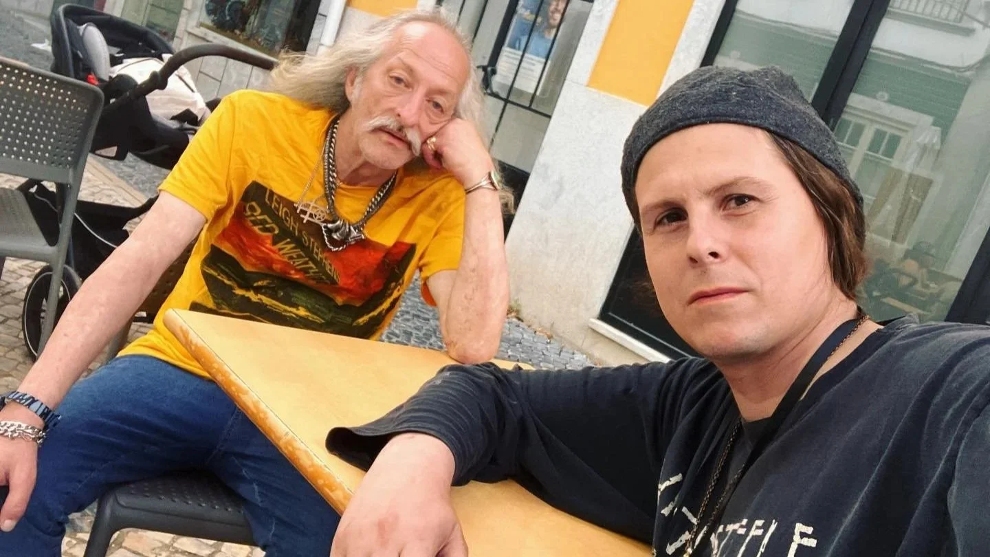 Two men sit at an outdoor cafe table. The older man on the left with long gray hair and mustache wearing a yellow graphic t-shirt leans his head on his hand, looking tired or bored. The younger man on the right with brown hair under a gray beanie and wearing a black shirt looks at the camera with a serious expression.