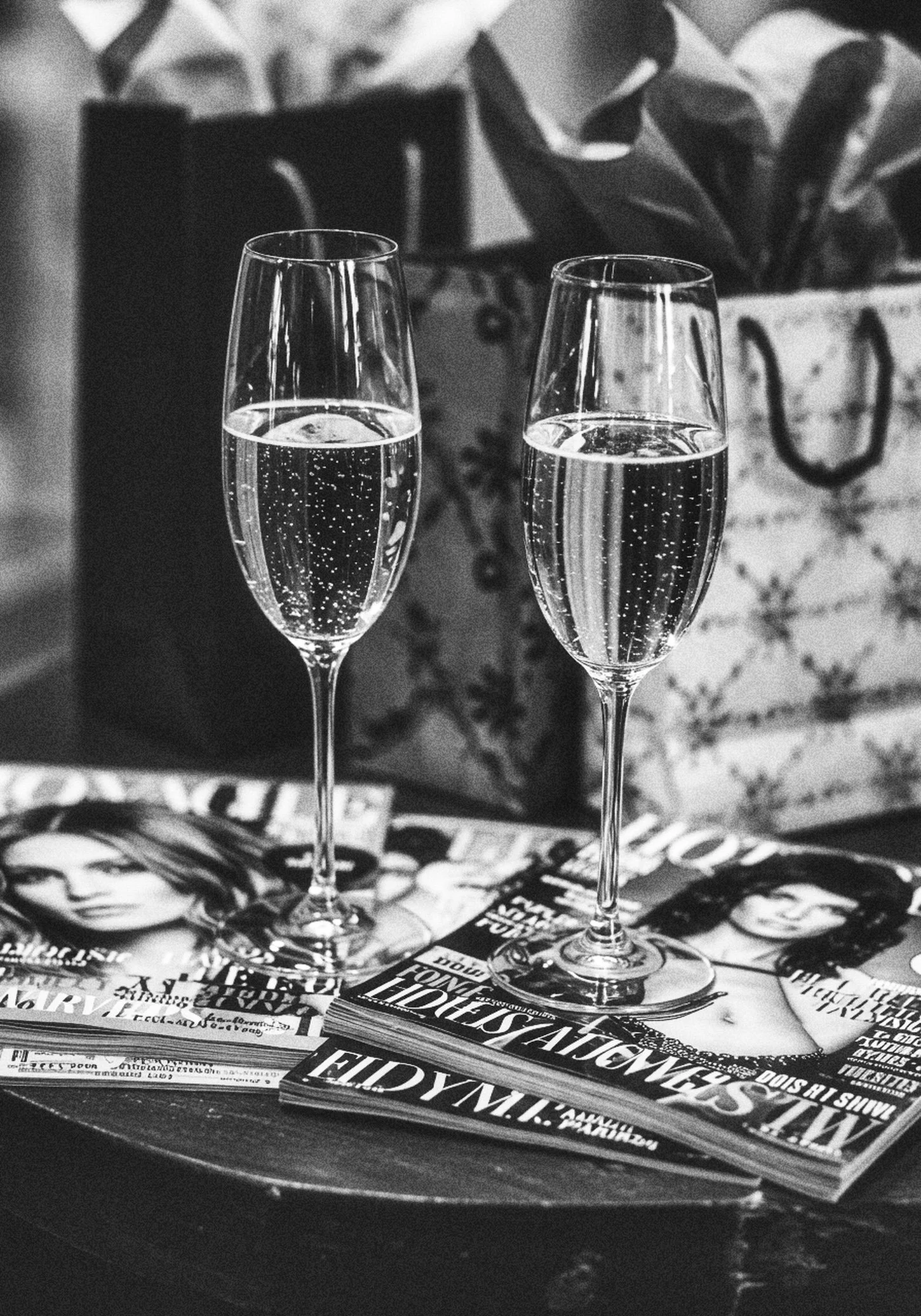 Two champagne glasses filled with sparkling wine are placed on top of magazines and newspapers on a table. There is a blurred background with a plant and a bag.