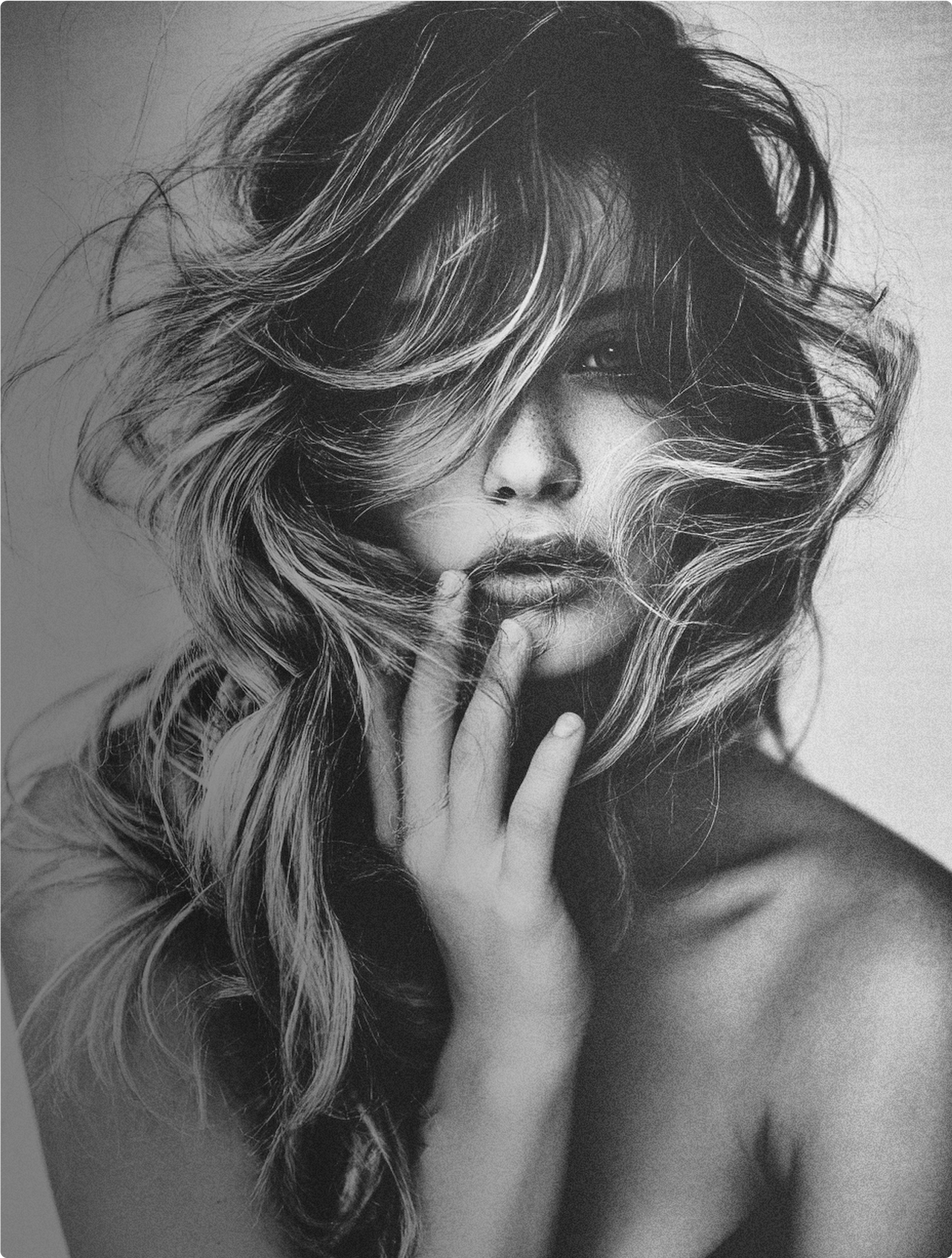 Black and white portrait of a woman with long, wavy hair covering part of her face, holding her finger to her lips.