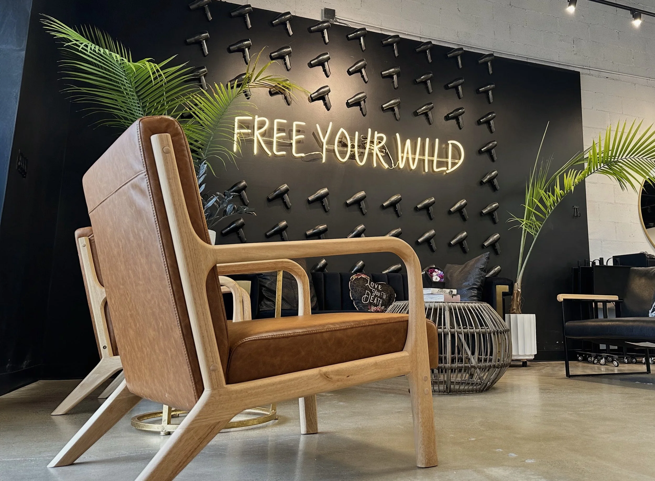 A black wall with multiple black hairdryers mounted on it, and a neon sign that says "FREE YOUR WILD". There are green plants and seating in front of the wall.