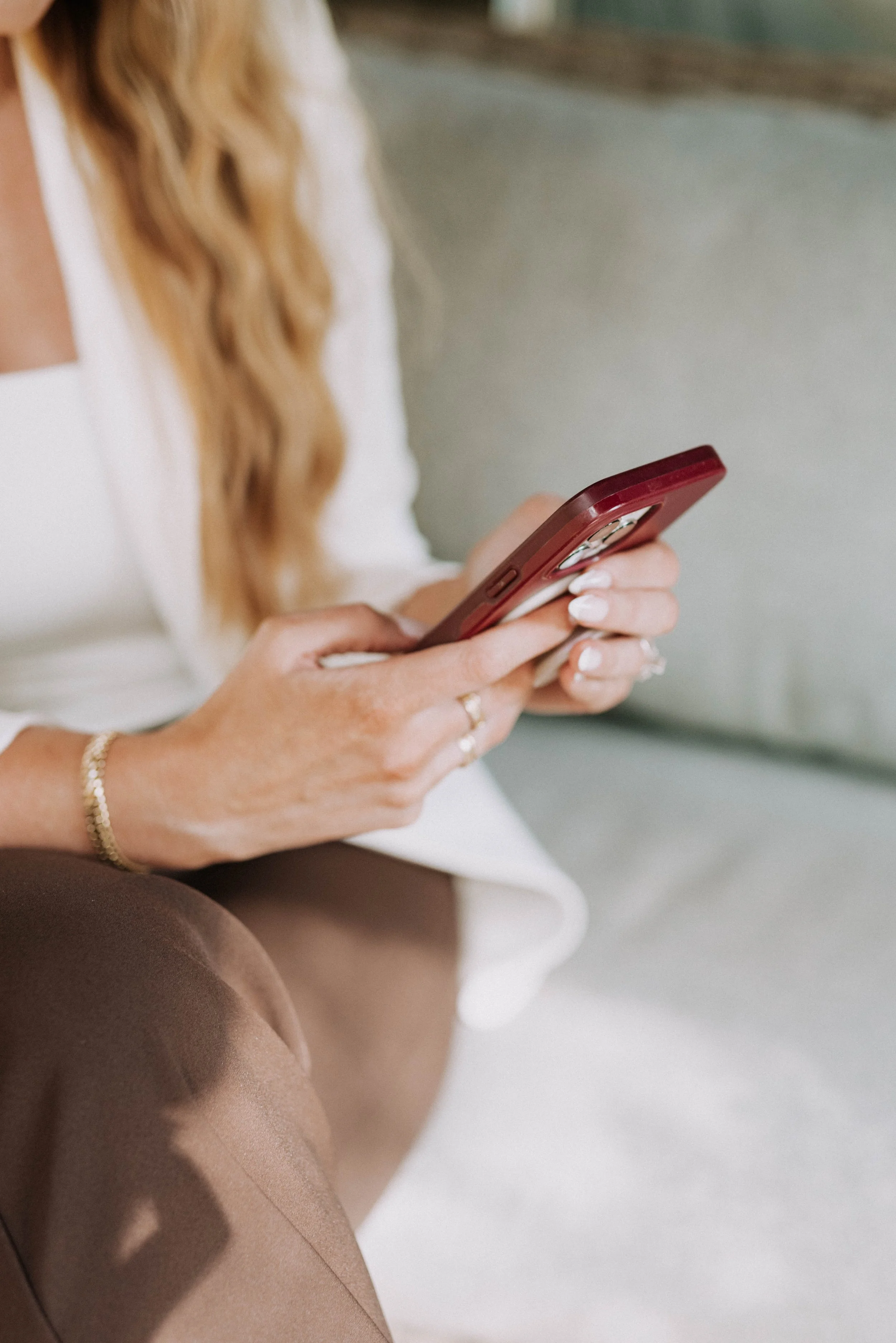 A woman sitting on a light-colored couch, using a red smartphone. She has long, wavy blond hair and is wearing a white top and brown pants, with jewelry on her wrist and fingers she is using her phone to manage social media for small businesses.