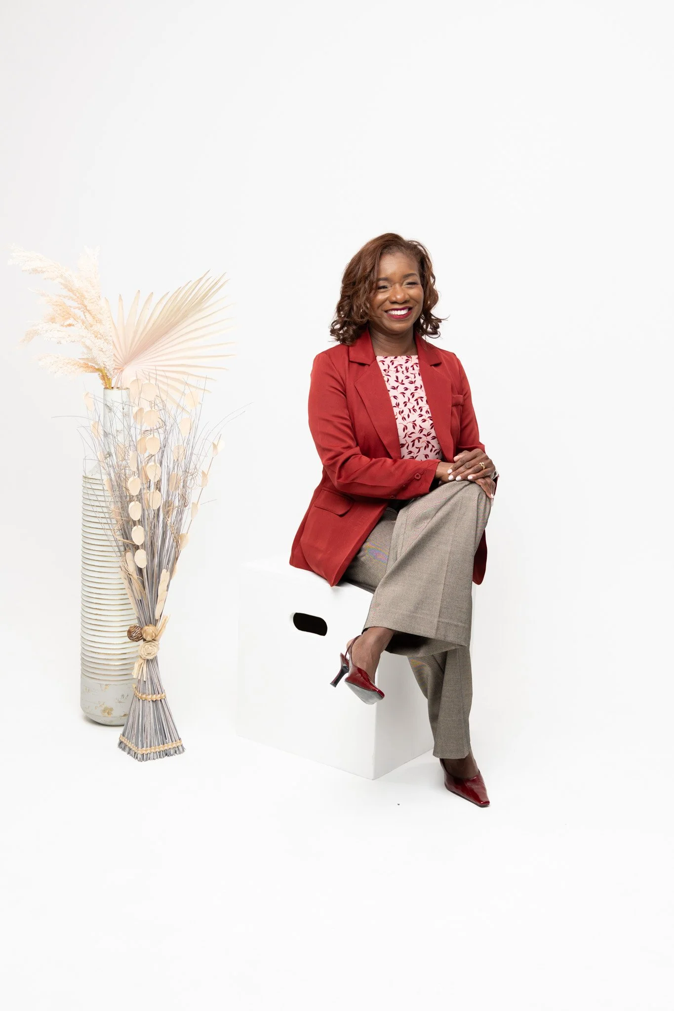 A smiling woman in a red blazer and beige wide-leg trousers seated on a white block. She has curly brown hair and is wearing red high-heeled shoes. To her left, there is a tall vase with pampas grass and decorative dried plants set against a white background.