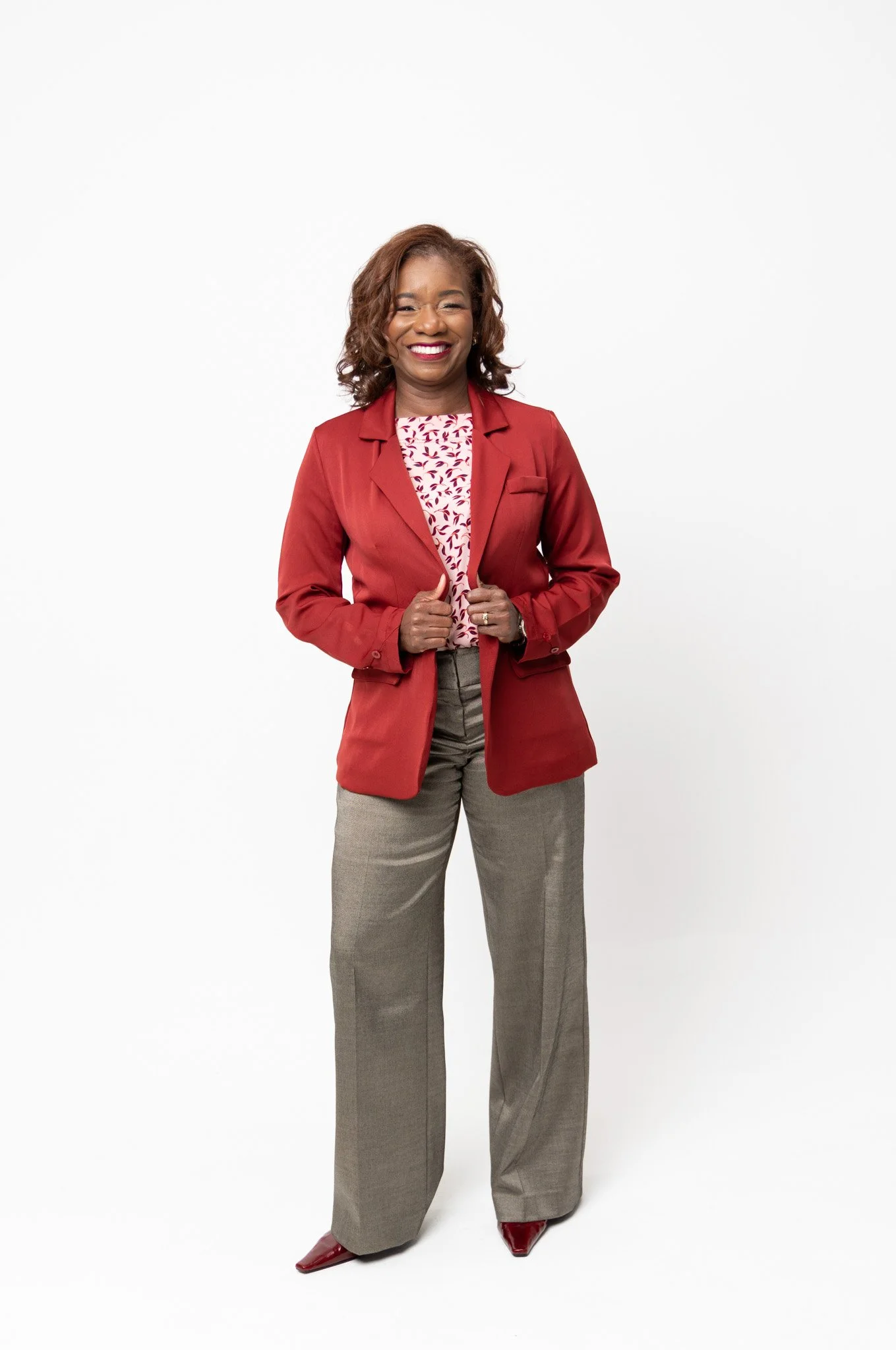 A smiling woman with medium-length curly hair wearing a red blazer, a patterned blouse, gray trousers, and red shoes, standing against a plain white background.