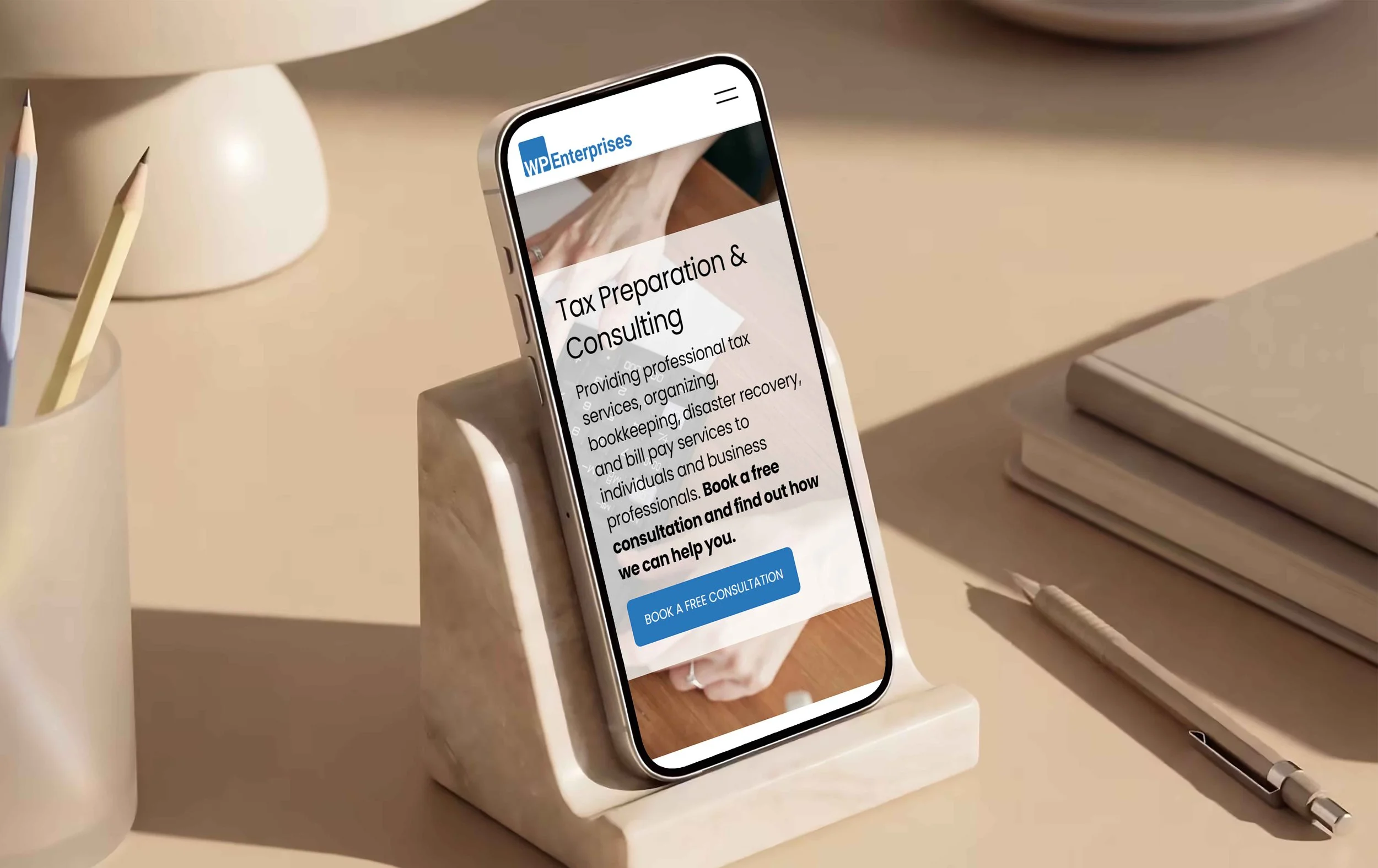 Smartphone on a stand displays a website for WP Enterprises, placed on a beige desk with notebooks, a pen, and pencils nearby.