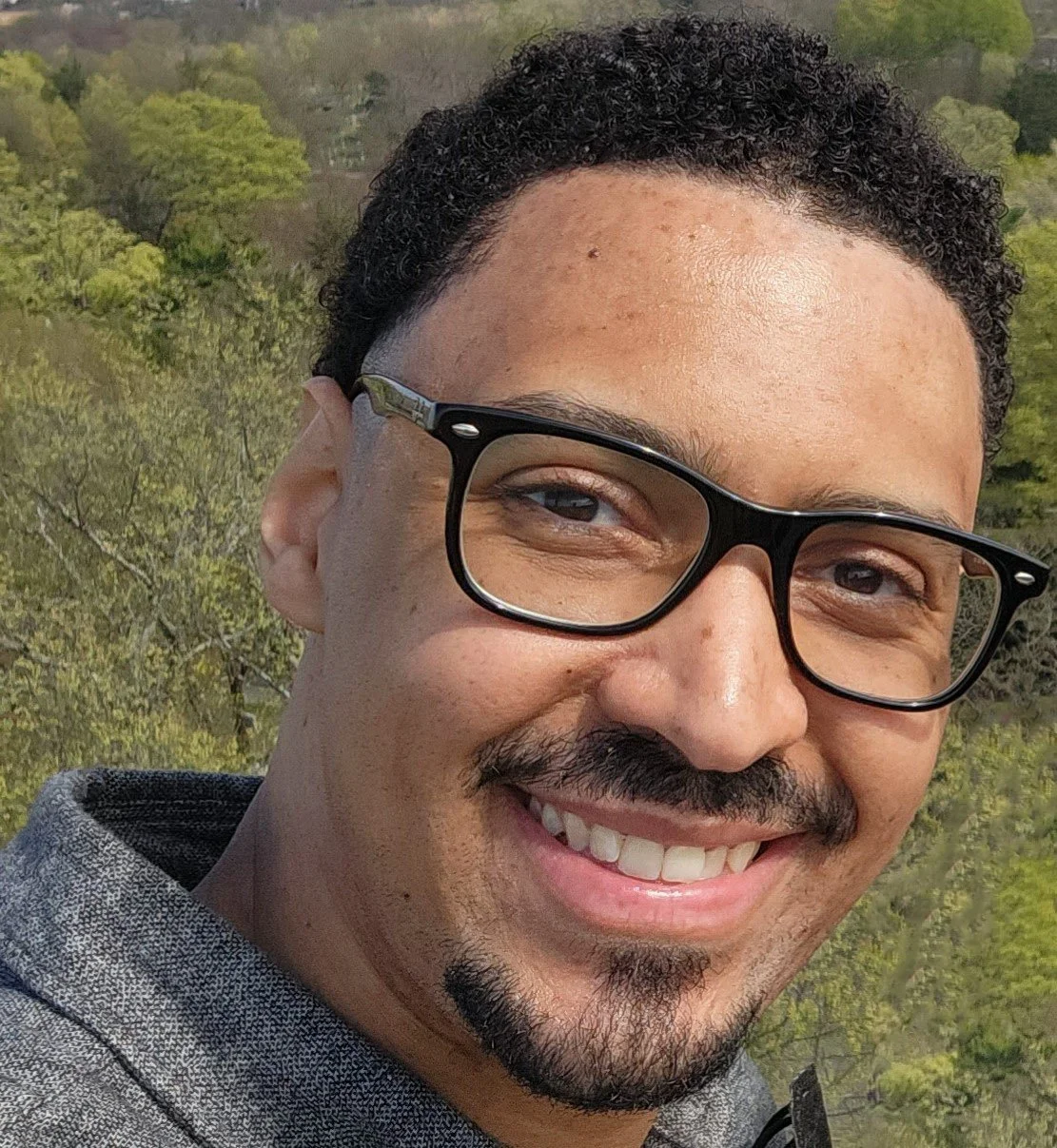 Close-up of a smiling man with glasses, short curly hair, and facial hair outdoors with green trees in the background.