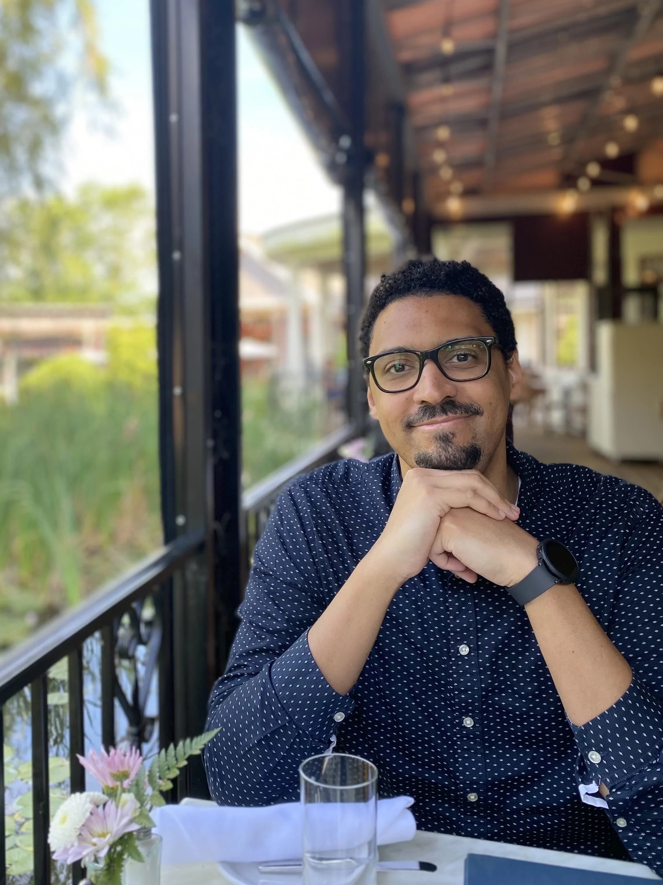 A man with glasses sitting at a table in a restaurant, smiling, with his chin resting on his hands, wearing a navy blue shirt with white polka dots and a black watch, in a bright outdoor setting with green foliage outside.