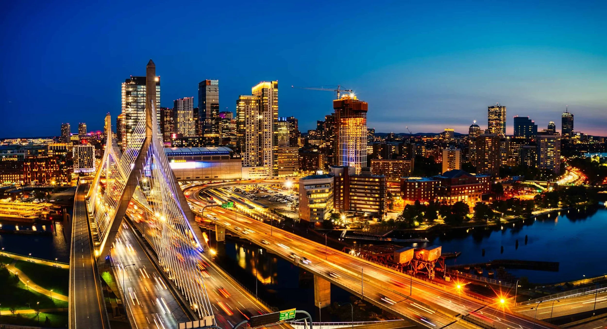 City skyline at dusk with illuminated skyscrapers, a cable-stayed bridge in the foreground, and a river reflecting city lights.