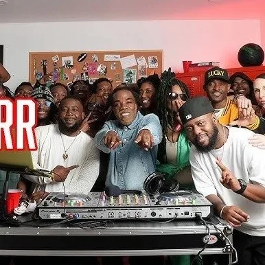 A group of diverse people celebrating around a DJ setup, smiling and making gestures in an indoor event space.