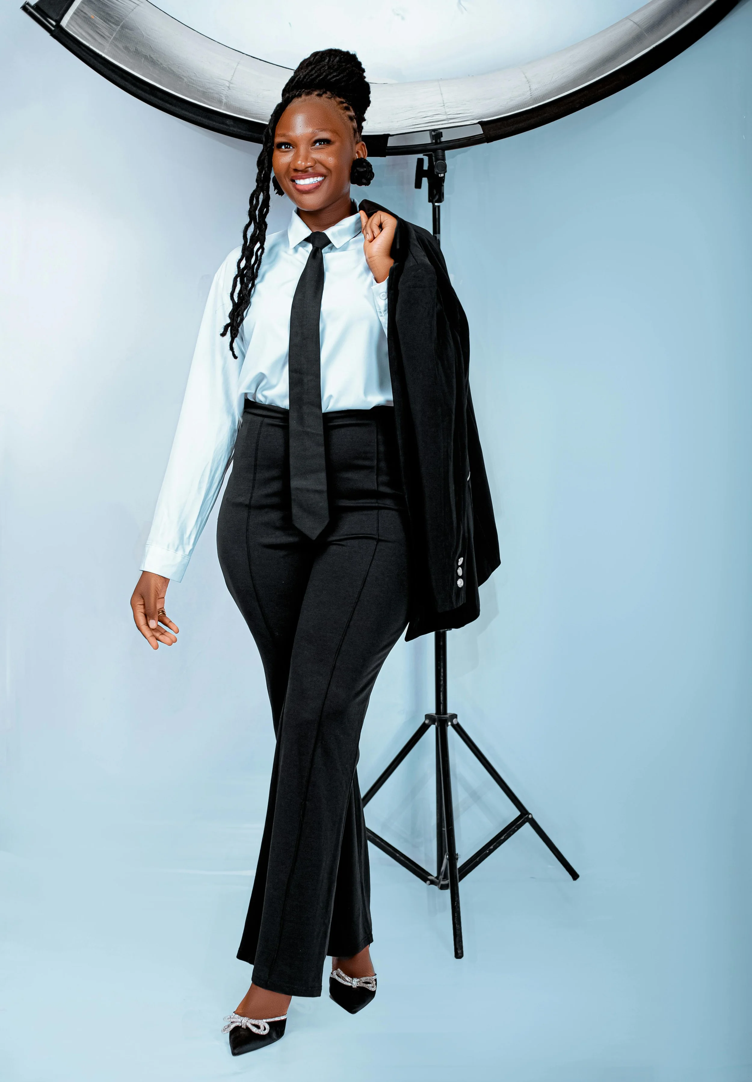 A woman dressed in professional attire standing in front of photography lighting equipment, holding a black blazer over her shoulder, smiling, wearing black high heels with silver embellishments, in a studio setting with a light blue background.