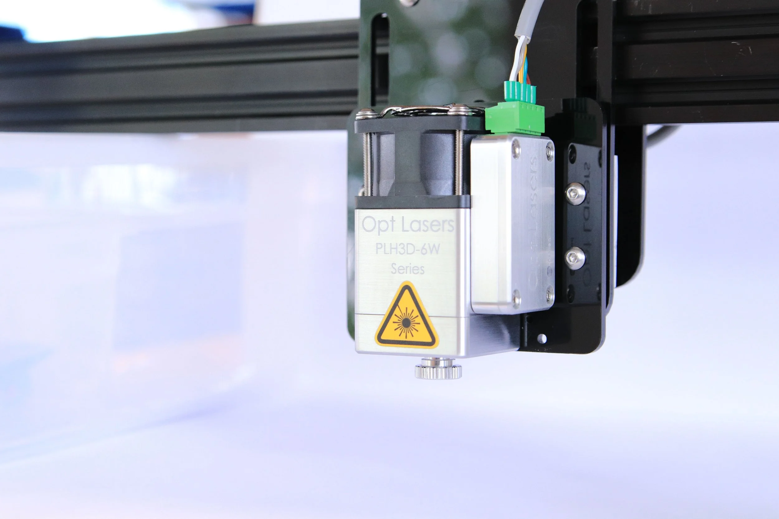 A close-up view of a laser module labeled 'Opt Lasers PLH3D-6W Series,' mounted on a black frame with a green connector and wires attached, and a yellow laser warning symbol on the front.