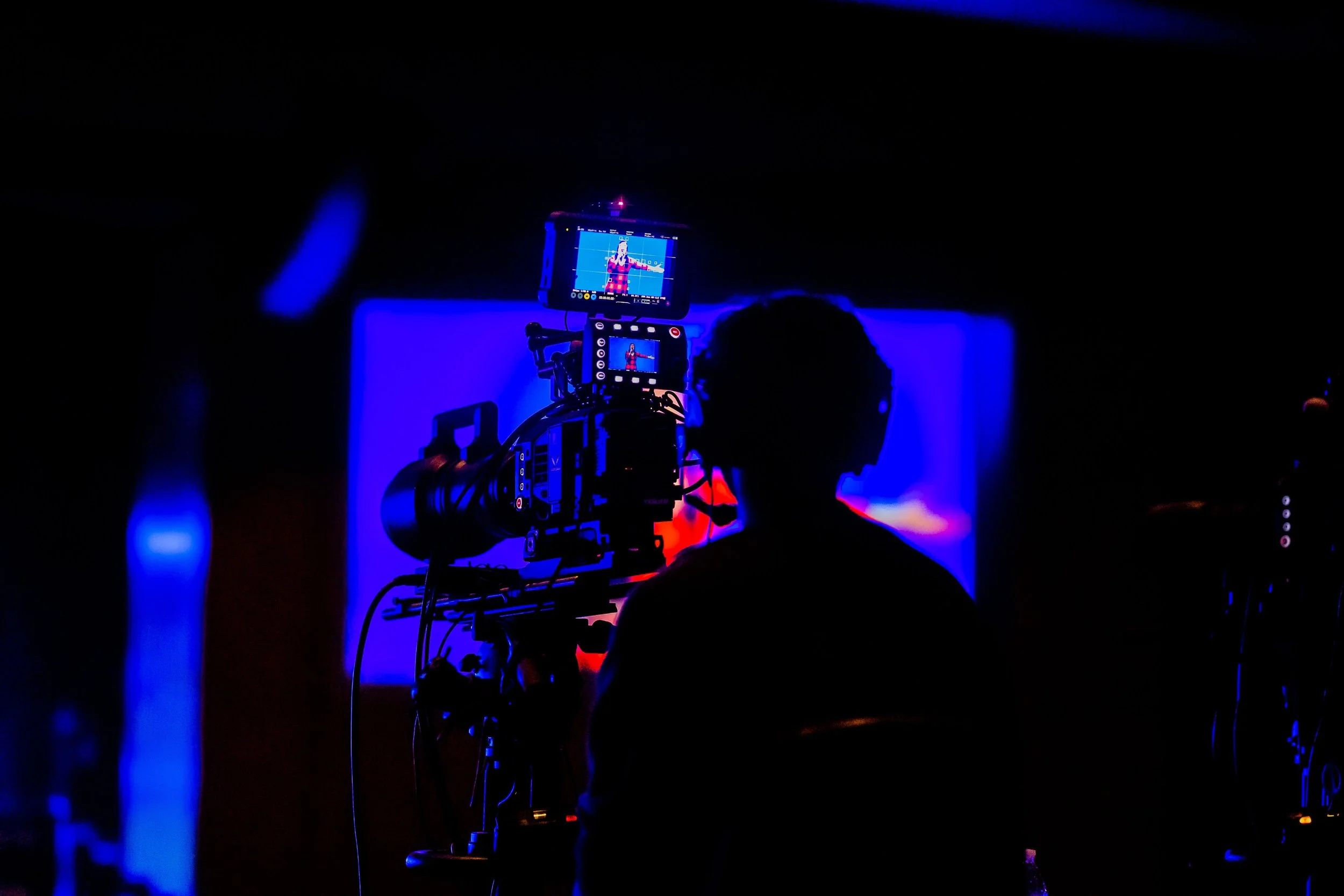 Camera filming a woman in a red checkered dress on stage with blue lighting.