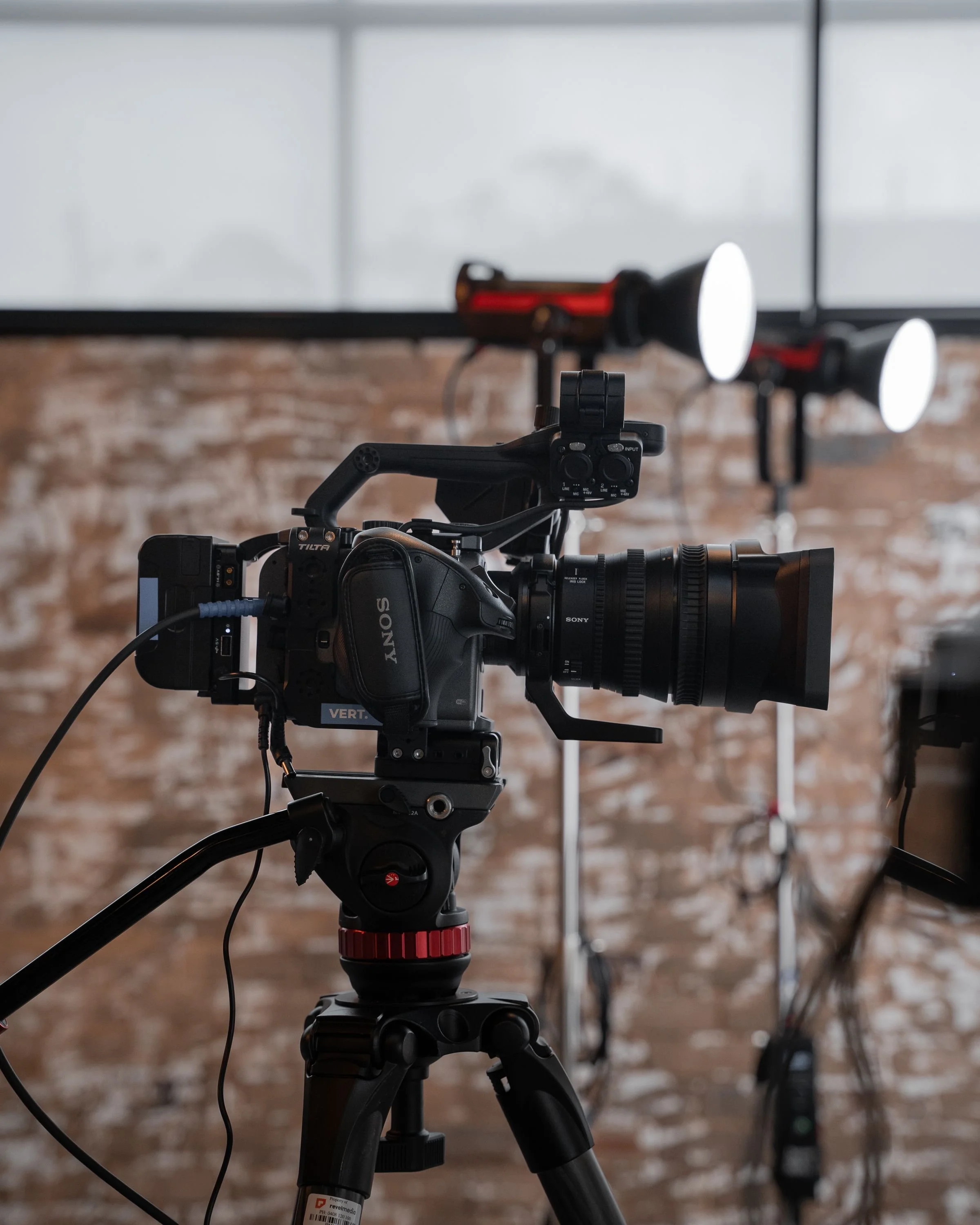 Professional video camera mounted on a tripod in an indoor studio with brick wall background and lighting equipment in the background.