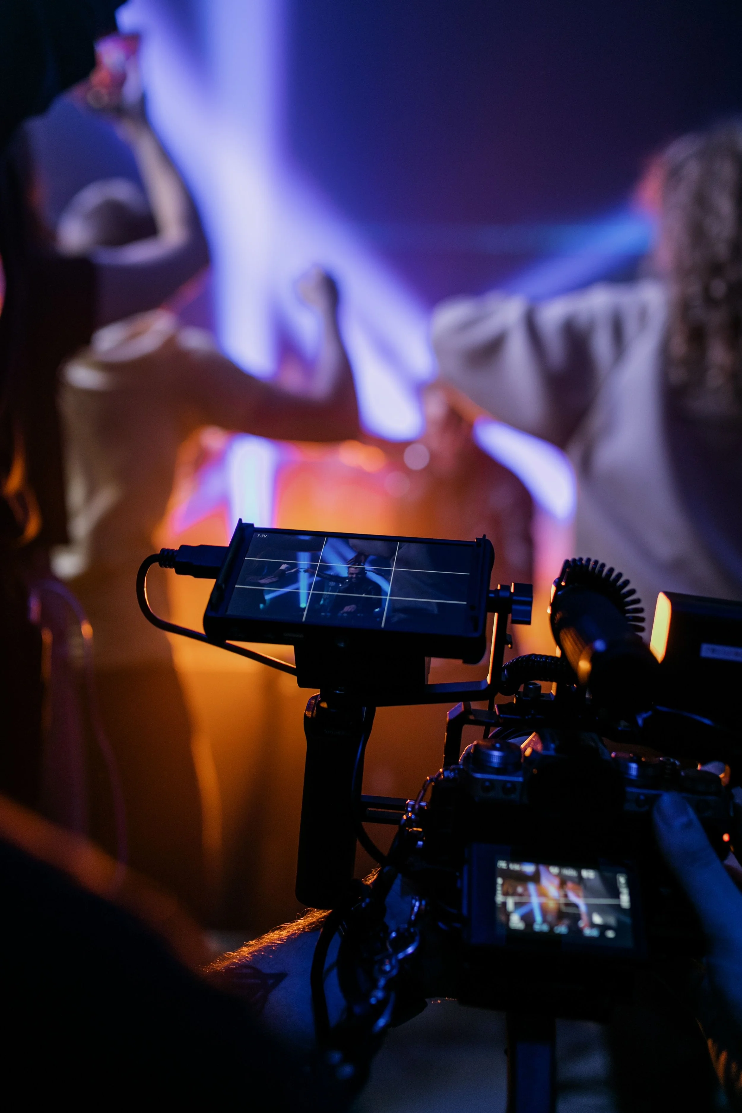 Behind-the-scenes view of film or video production setup with a camera and monitor in focus, with a person operating the equipment and two people in the background waving or gesturing in a dimly lit environment with blue and orange lighting.