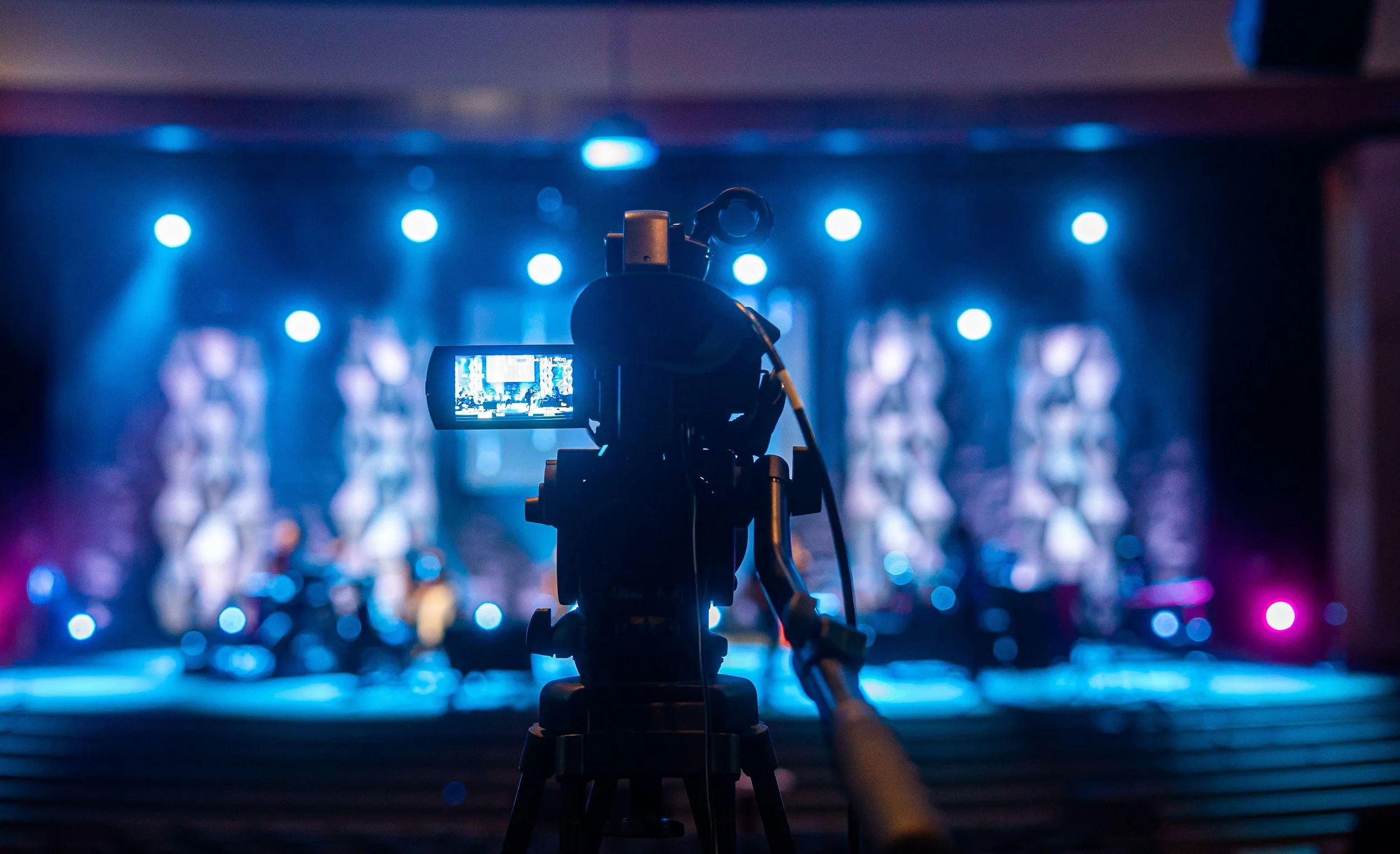 Close-up of a professional video camera on a tripod filming a stage with colorful lights in the background.