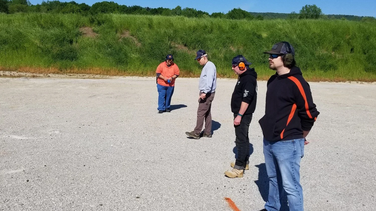Four people wearing hearing protection and safety glasses stand outdoors on a gravel or dirt area, with a grassy hill in the background, under a clear blue sky.