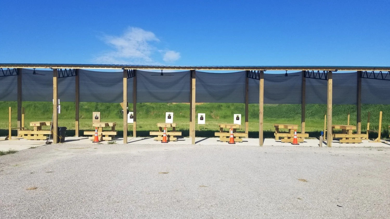 Row of outdoor shooting range lanes with targets, benches, and orange cones under a shaded structure, with grassy field and blue sky in the background.