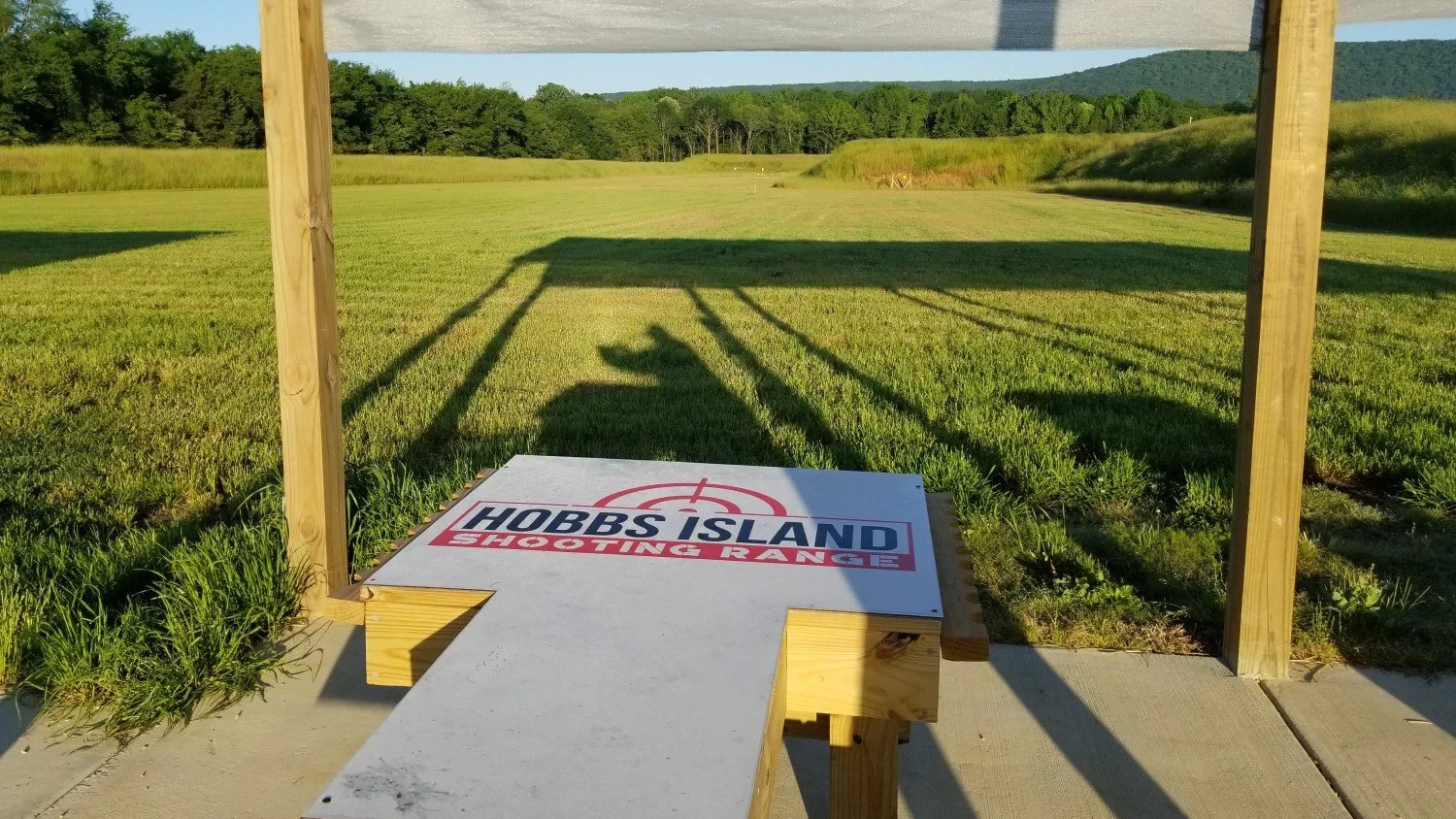A wooden shooting range platform with a sign that reads "Hobbs Island Shooting Range" situated on the ground in an open grassy area, with shadows cast by a wooden frame structure. In the background, there are green fields, trees, and distant hills under a clear sky.