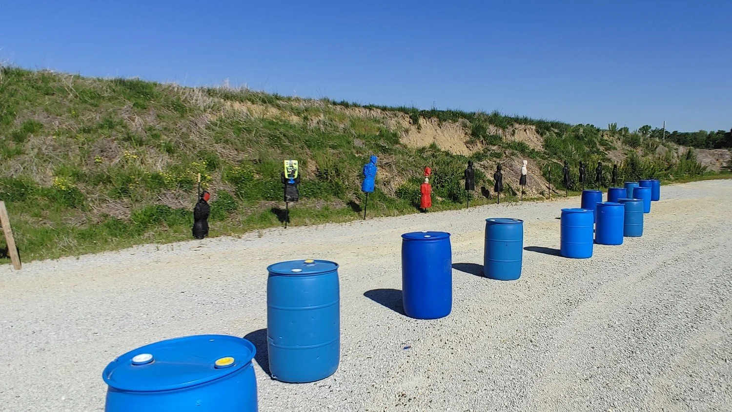 A row of blue barrels on a gravel surface in front of a grassy hillside with shooting targets dressed as people in various colors. The targets are spaced apart along the hillside.