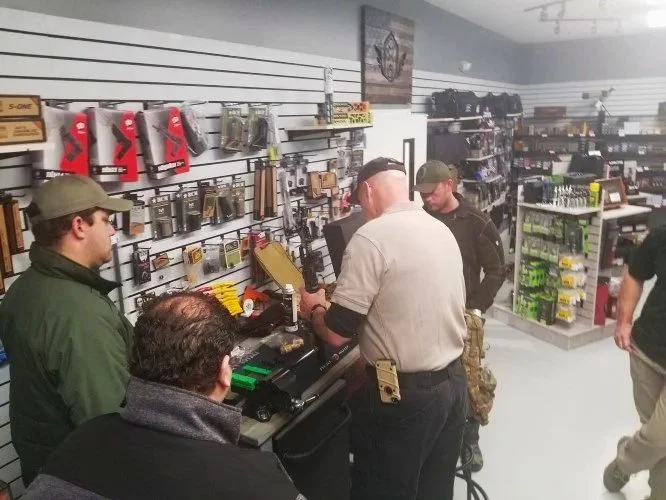 Four men are gathered around a checkout counter inside a store that sells outdoor, tactical, or sporting gear. The wall behind them displays various tools, accessories, and gear items. One man, wearing a gray polo shirt and baseball cap, is handling an item at the register, while the others observe.