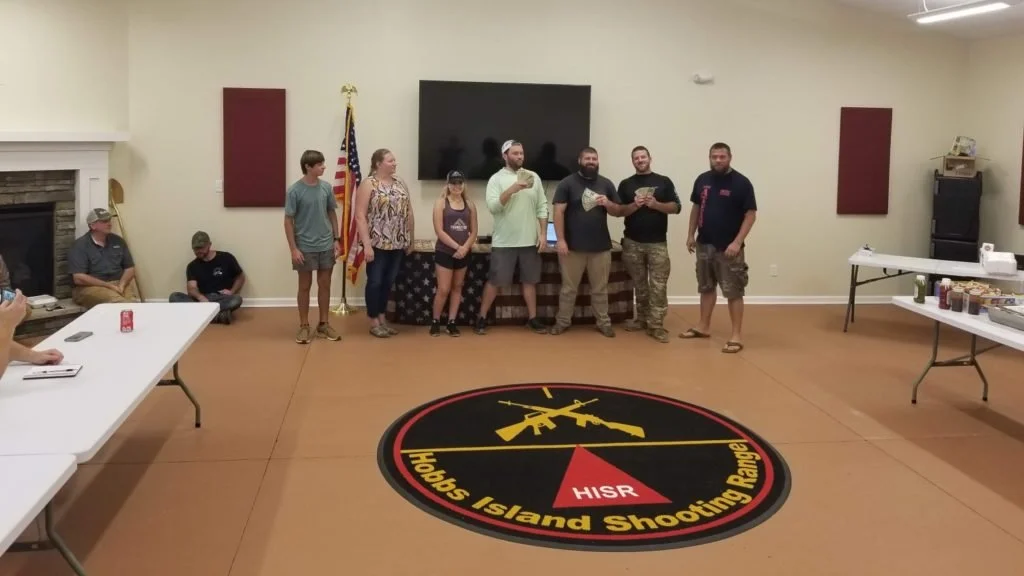 Group of people standing in a large open, training room with an American flag, a large TV, and a table with food and drinks, on a floor with a round logo that reads "Hobbs Island Shooting Range."