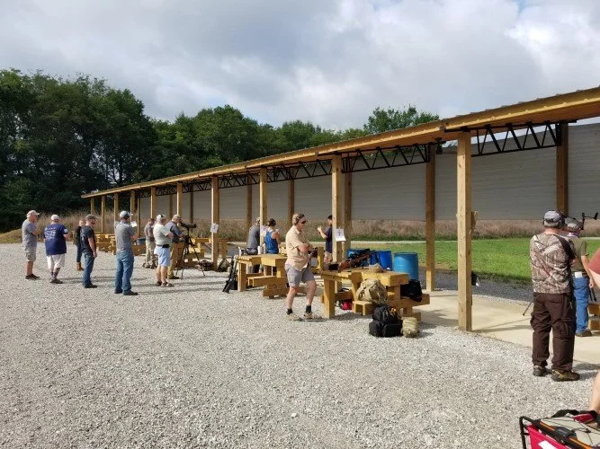 People participating in a shooting event at an outdoor shooting range, using rifles and pistols under a covered structure with targets in the background.