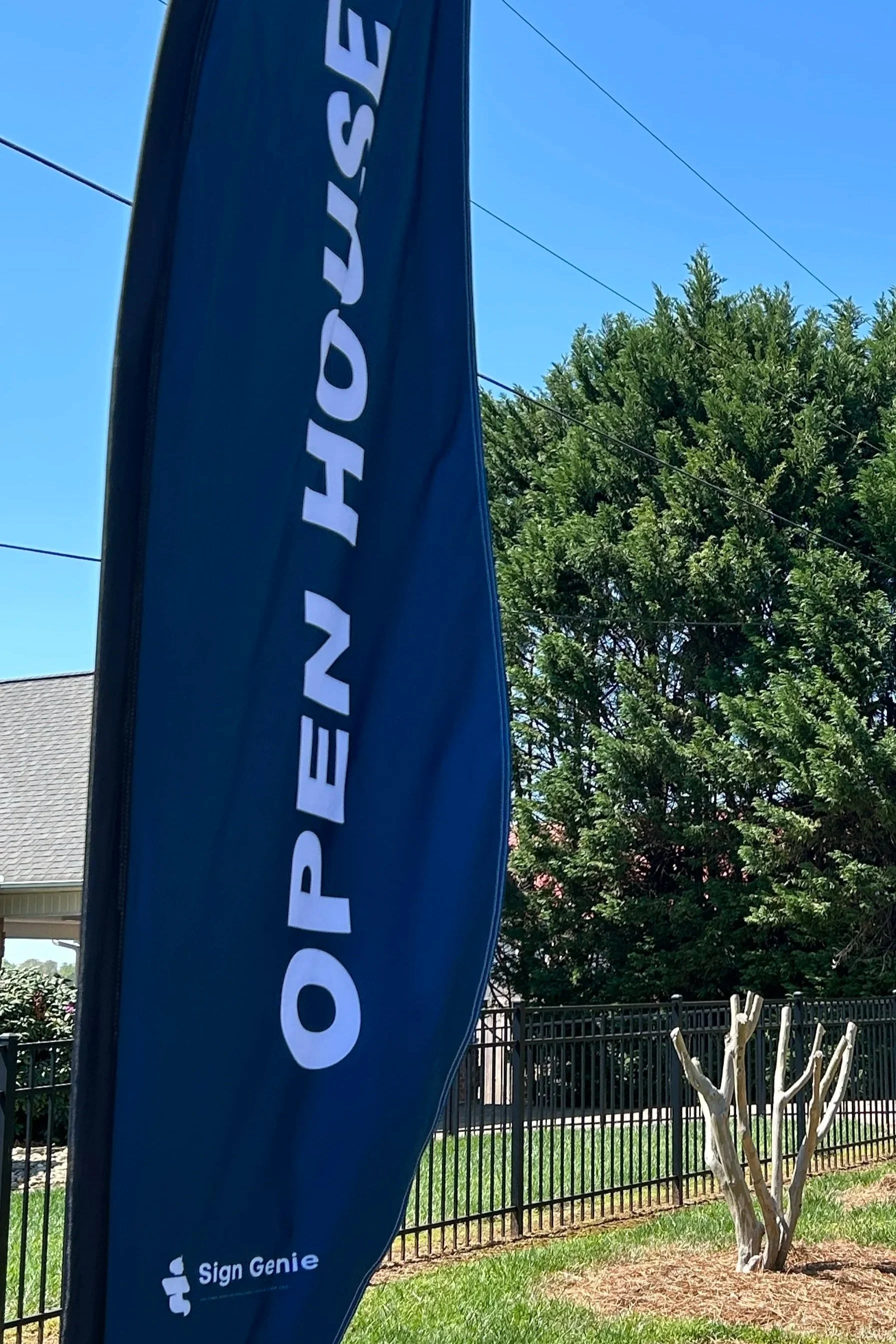Blue outdoor flag with white text stating "OPEN HOUSE" and a small logo for Sign Genie at the bottom, set in front of a fenced yard with trees and a house nearby.