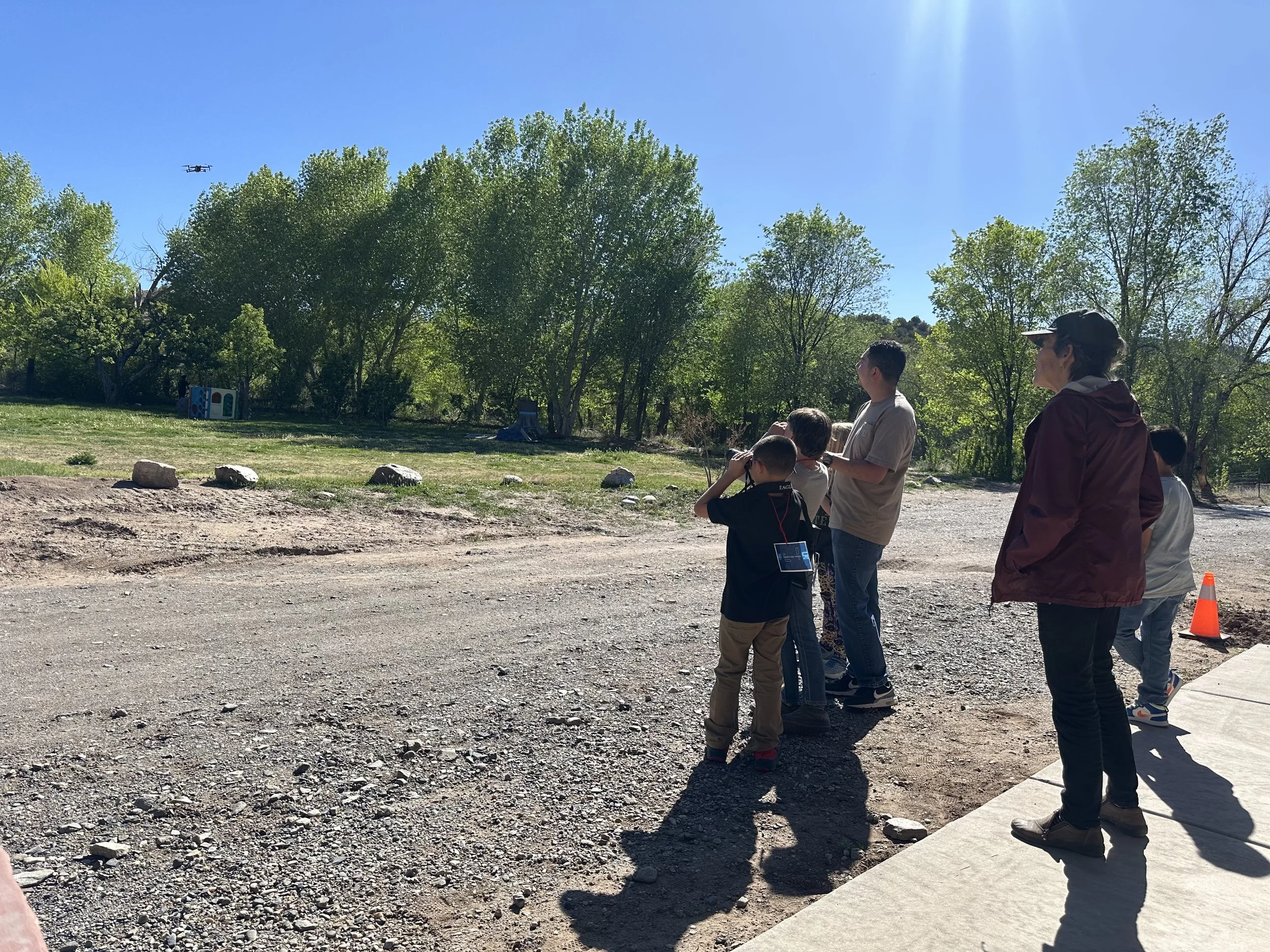 A group of children and an adult standing outdoors on a sunny day, observing a drone flying in the sky above a natural setting with trees.