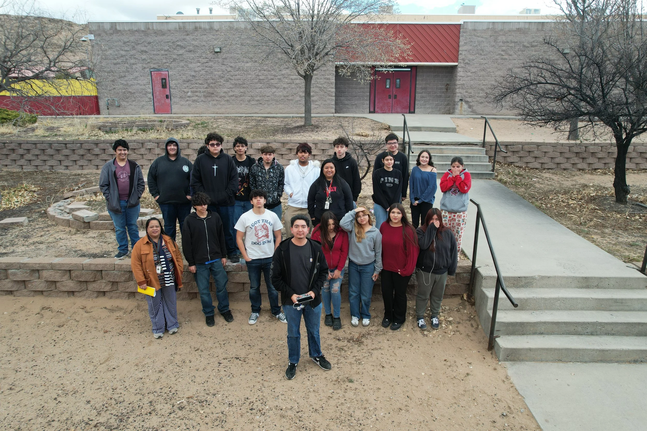 Group of diverse students and teachers standing outside near stairs in a schoolyard, with a brick building and leafless trees in the background.