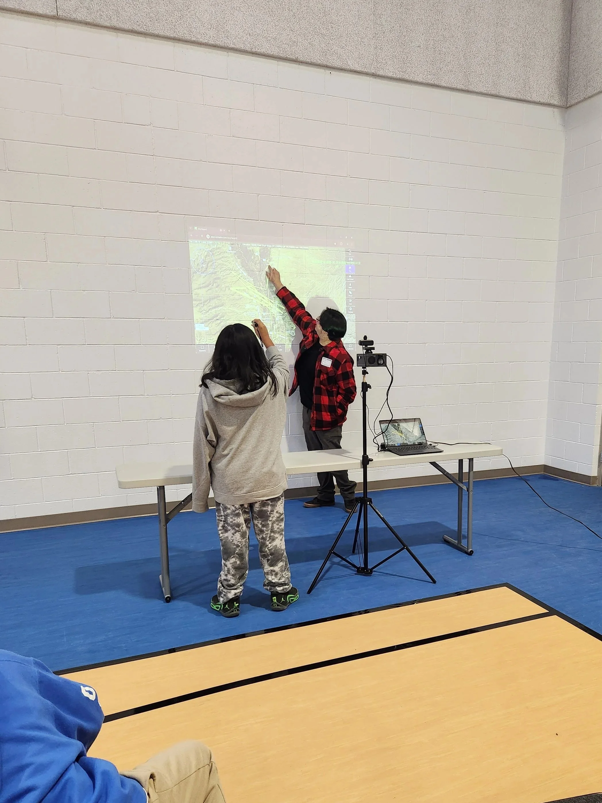A group of three people are in a gymnasium, one person is pointing at a projected map on the wall, and the other two are observing. One person is standing while the other is seated nearby. The map is displayed on a white brick wall, and there is technical equipment set up nearby.