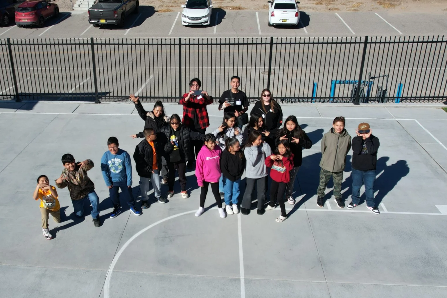 A group of children and a few adults on a basketball court posing for a photo. The children are standing in a line and making peace signs or pointing. The adults are standing behind the children, with one person taking a photo. The court has a metal fence and parked cars in the background.