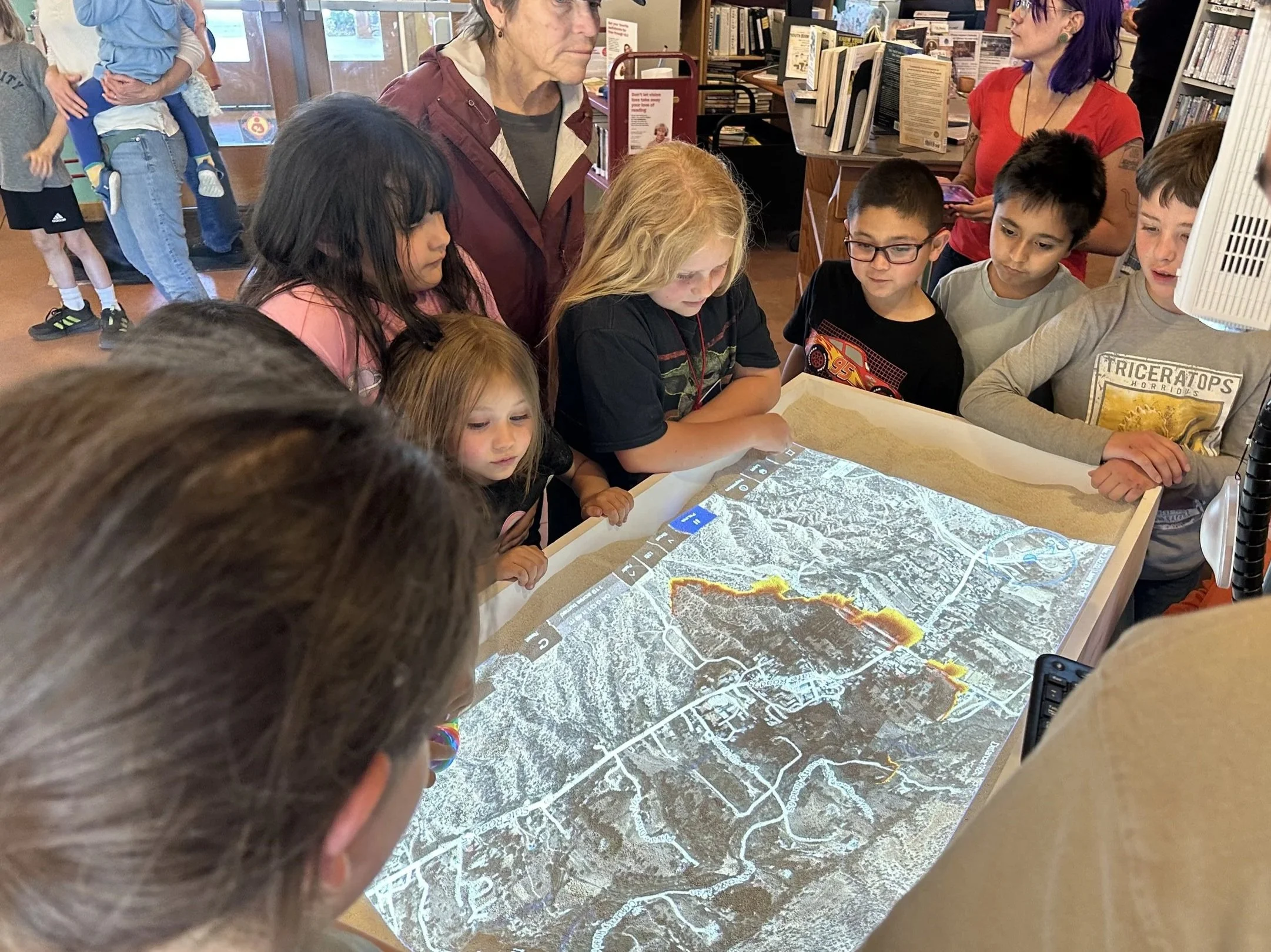 People gathered around a large map or terrain diagram on a table, inside a bookstore or library.