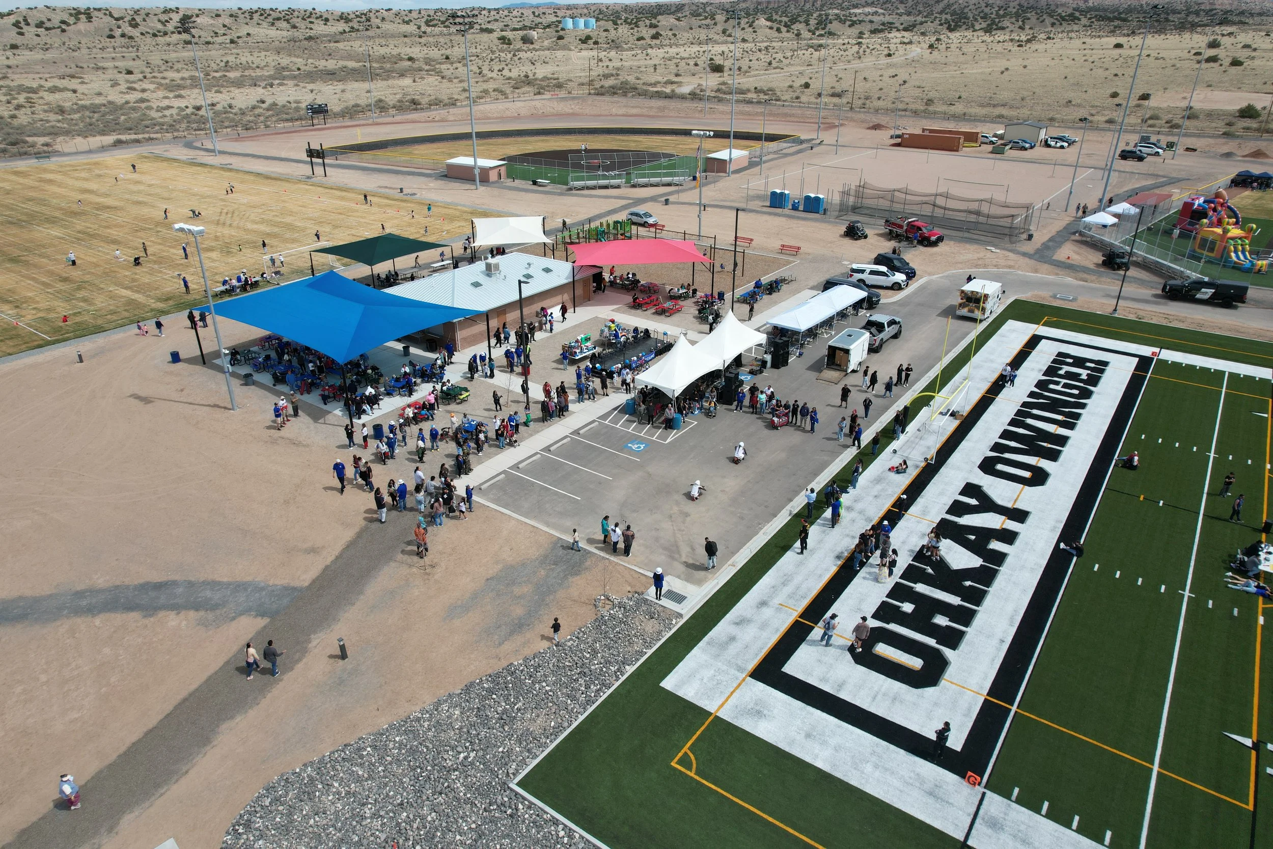 An aerial view of a sports complex featuring a football field, baseball field, and a playground, with people gathered around tents and parked cars in a desert landscape.