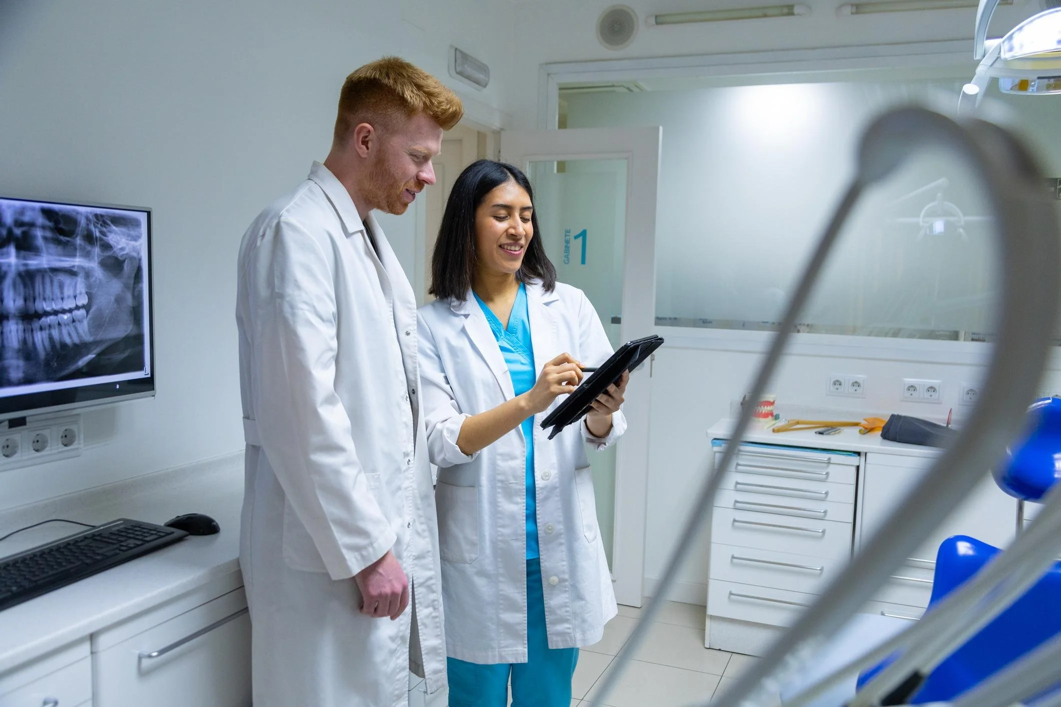 Two medical professionals, a man and a woman, are standing together in a hospital room, looking at a tablet device with an X-ray of a human skull on a monitor nearby.