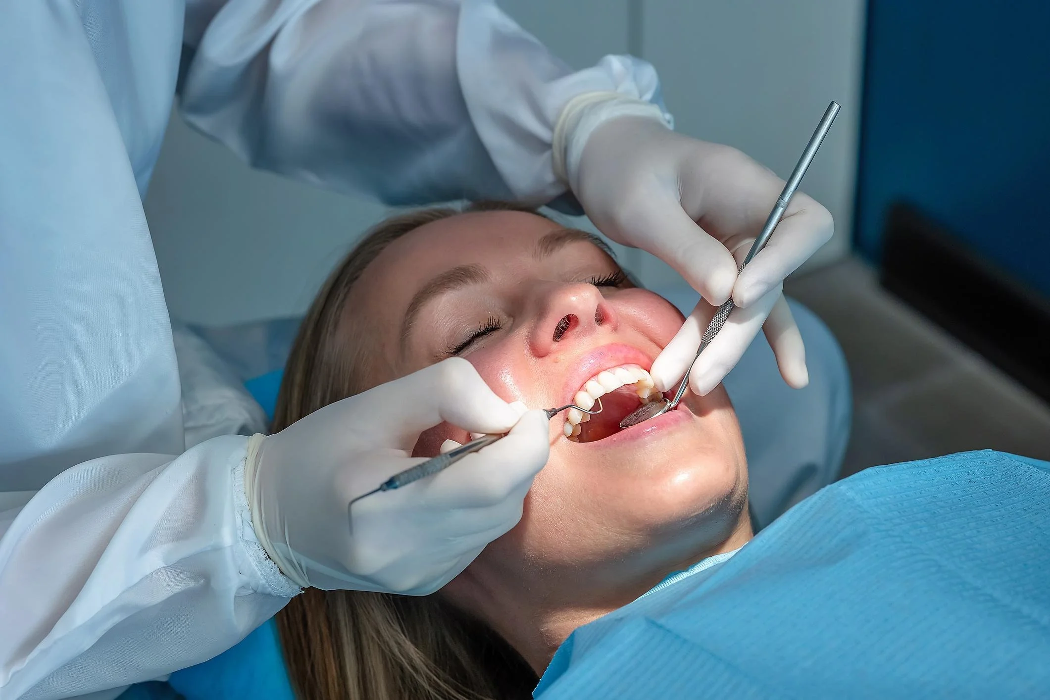 A woman lying back with closed eyes during a dental examination as a dentist in gloves uses dental tools in her mouth.