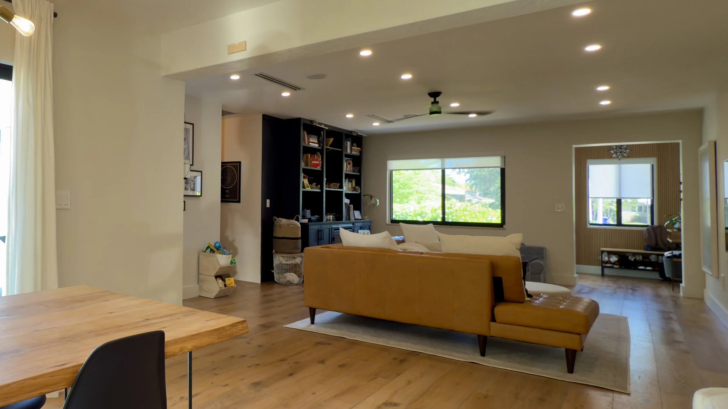 Living room with a yellow sofa, wooden dining table and chairs, black bookshelf, large windows, and wooden flooring.