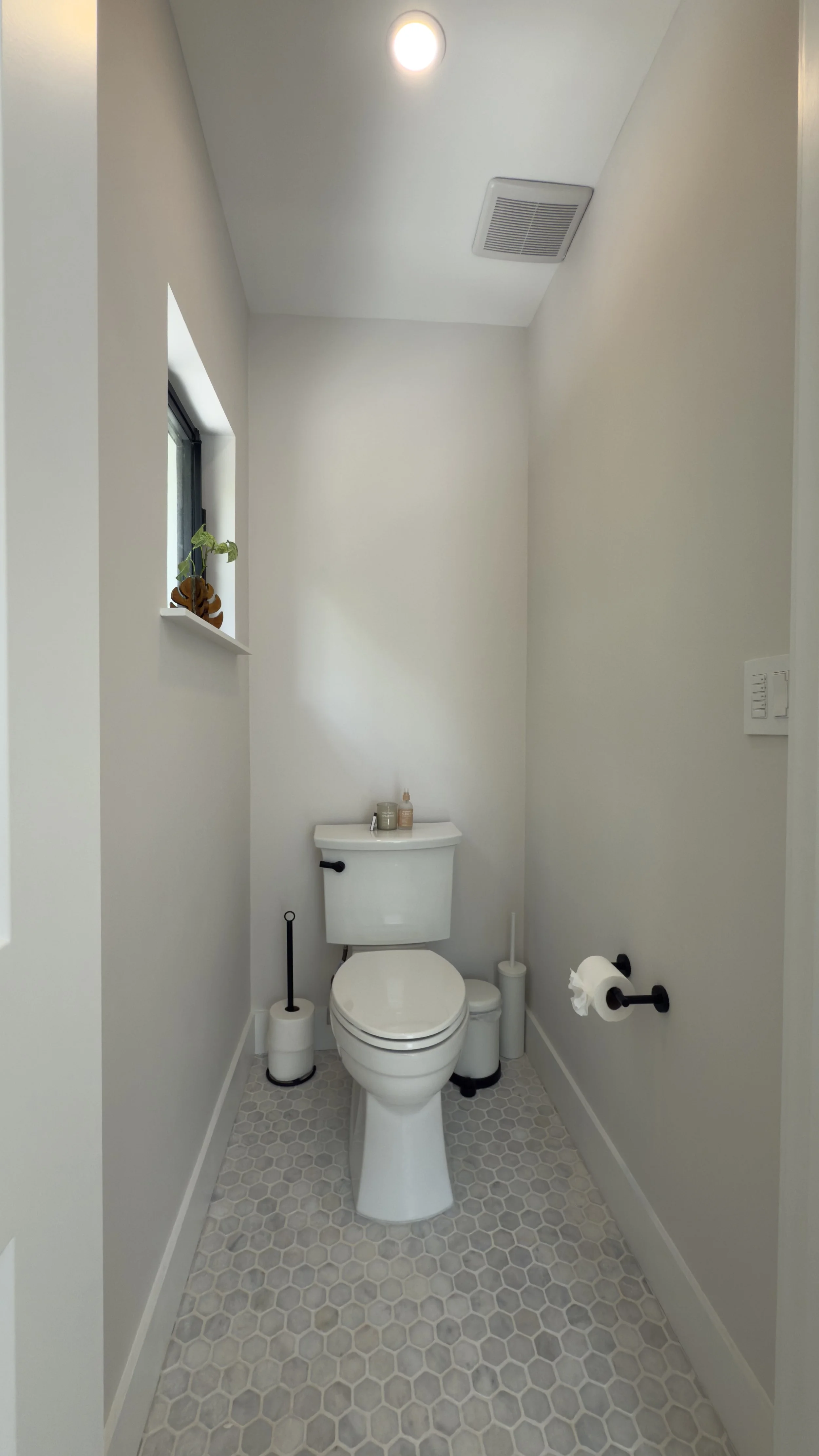 A small, minimalist bathroom with a white toilet, light gray hexagonal tile floor, and a small window with a plant. The walls are painted white, and there is a toilet paper roll on a black holder, along with a toilet brush and some small bottles on t