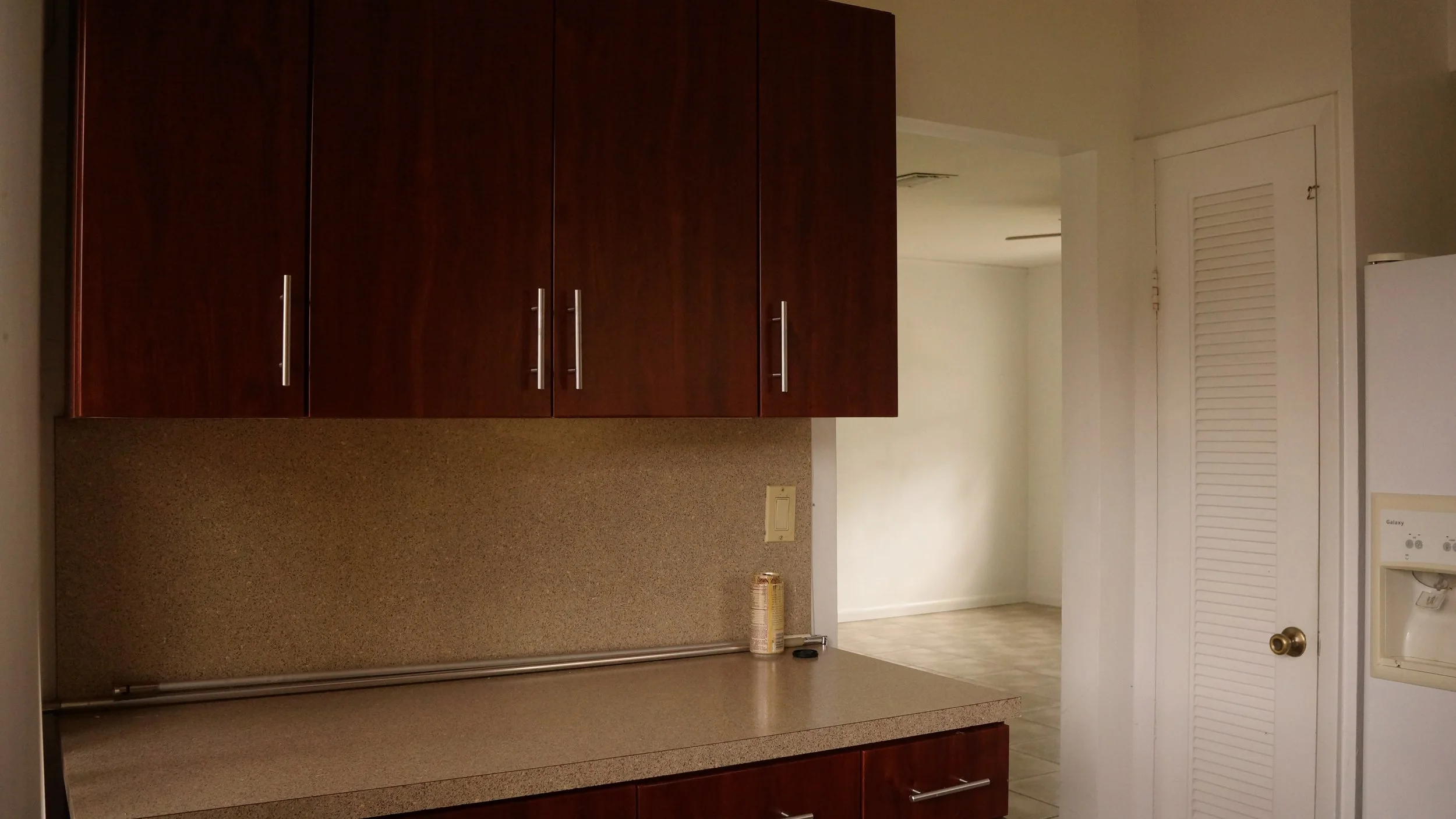 A kitchen with dark wood cabinets, a beige countertop, and a beige textured backsplash. There is a small can and a remote on the counter, with a white refrigerator visible on the right. A door with louvered panels is seen to the right, leading to ano