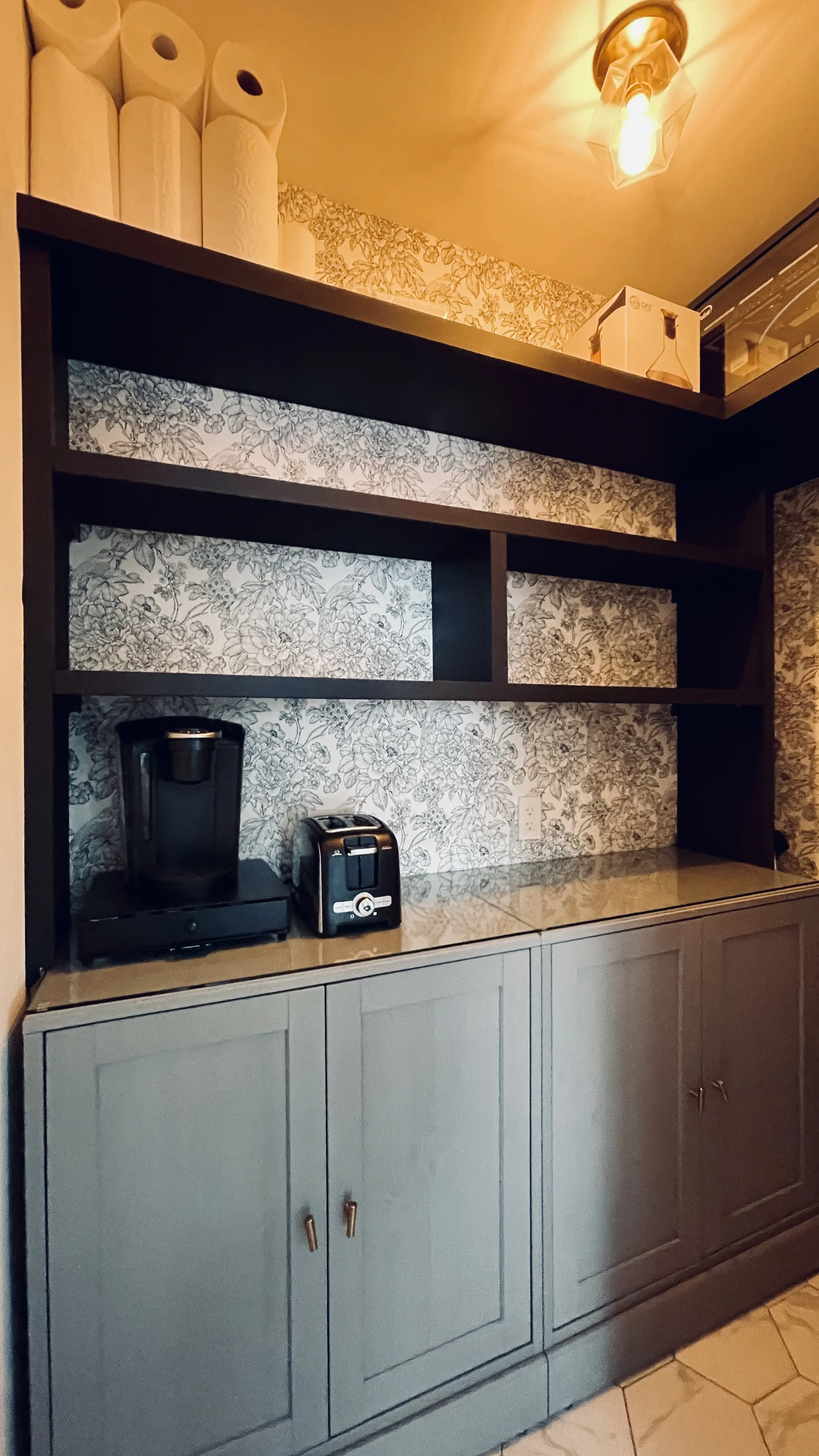 Kitchen cabinet with coffee maker, toaster, and appliances on the countertop, floral wallpaper behind, and a ceiling light above.