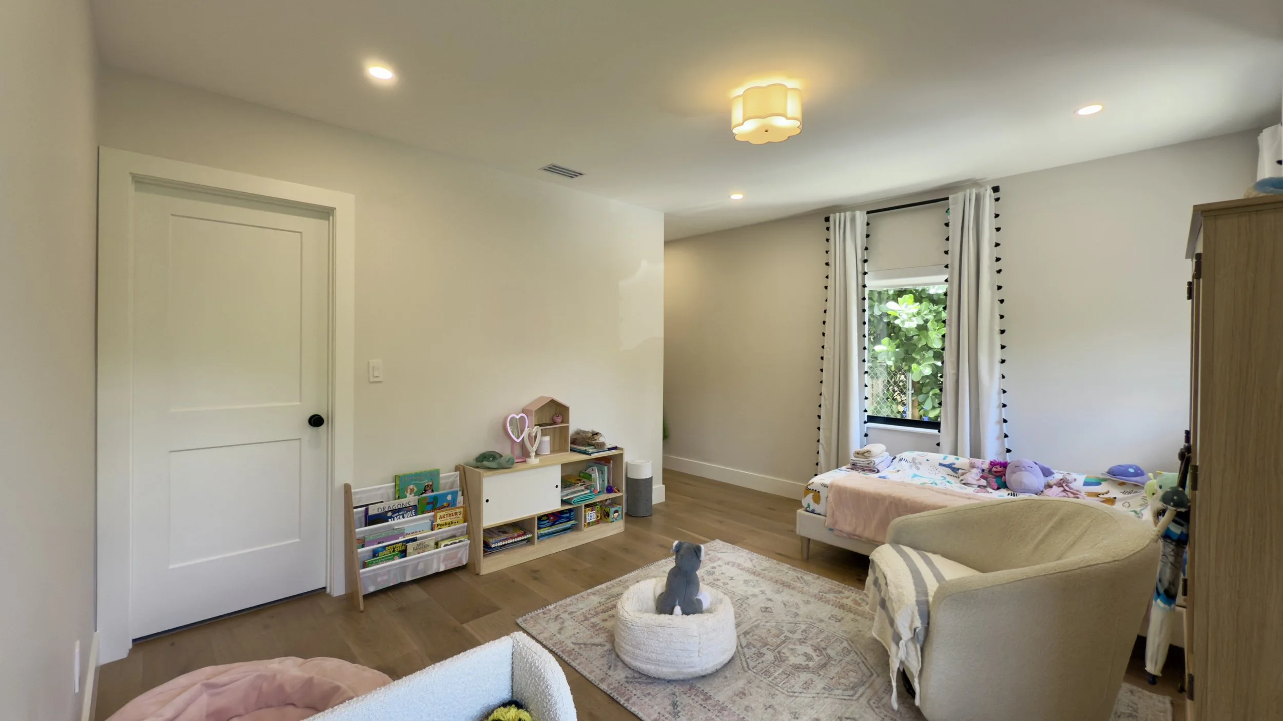 A cozy children's bedroom with a bed, armchair, bookshelf, and a stuffed animal on a rug near the window with leafy greenery outside.