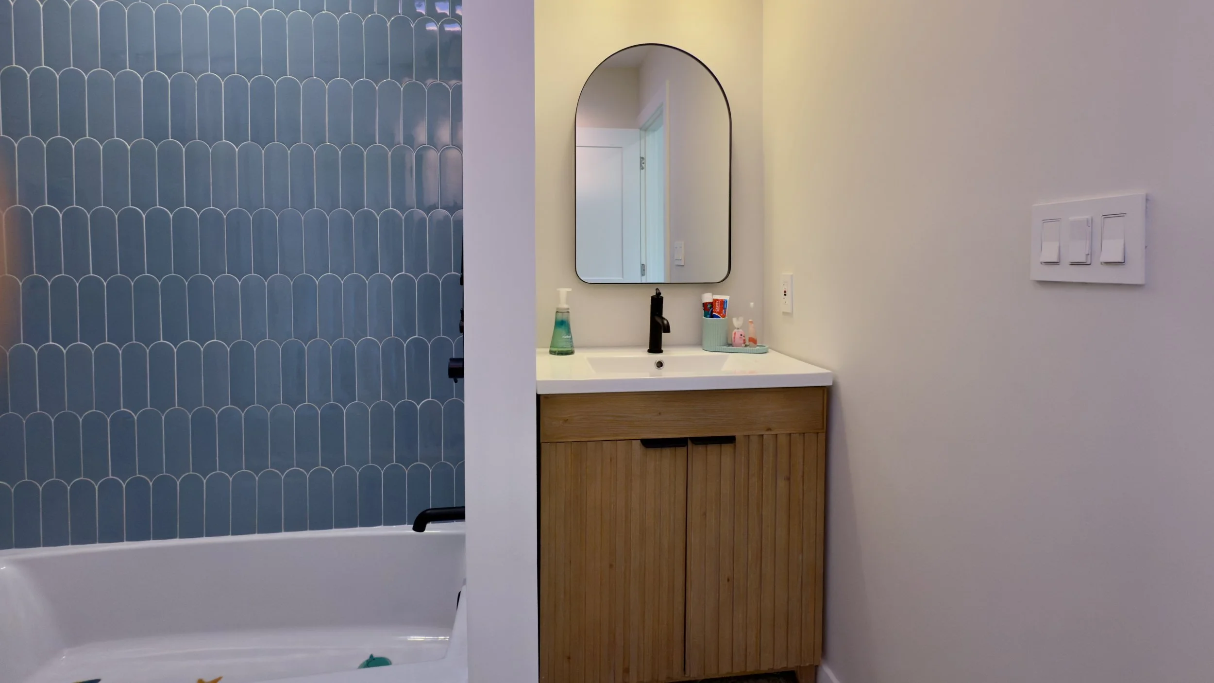 Bathroom with wooden vanity, oval mirror, and a sink with soap dispenser. Blue tiled shower behind a white wall on the left.
