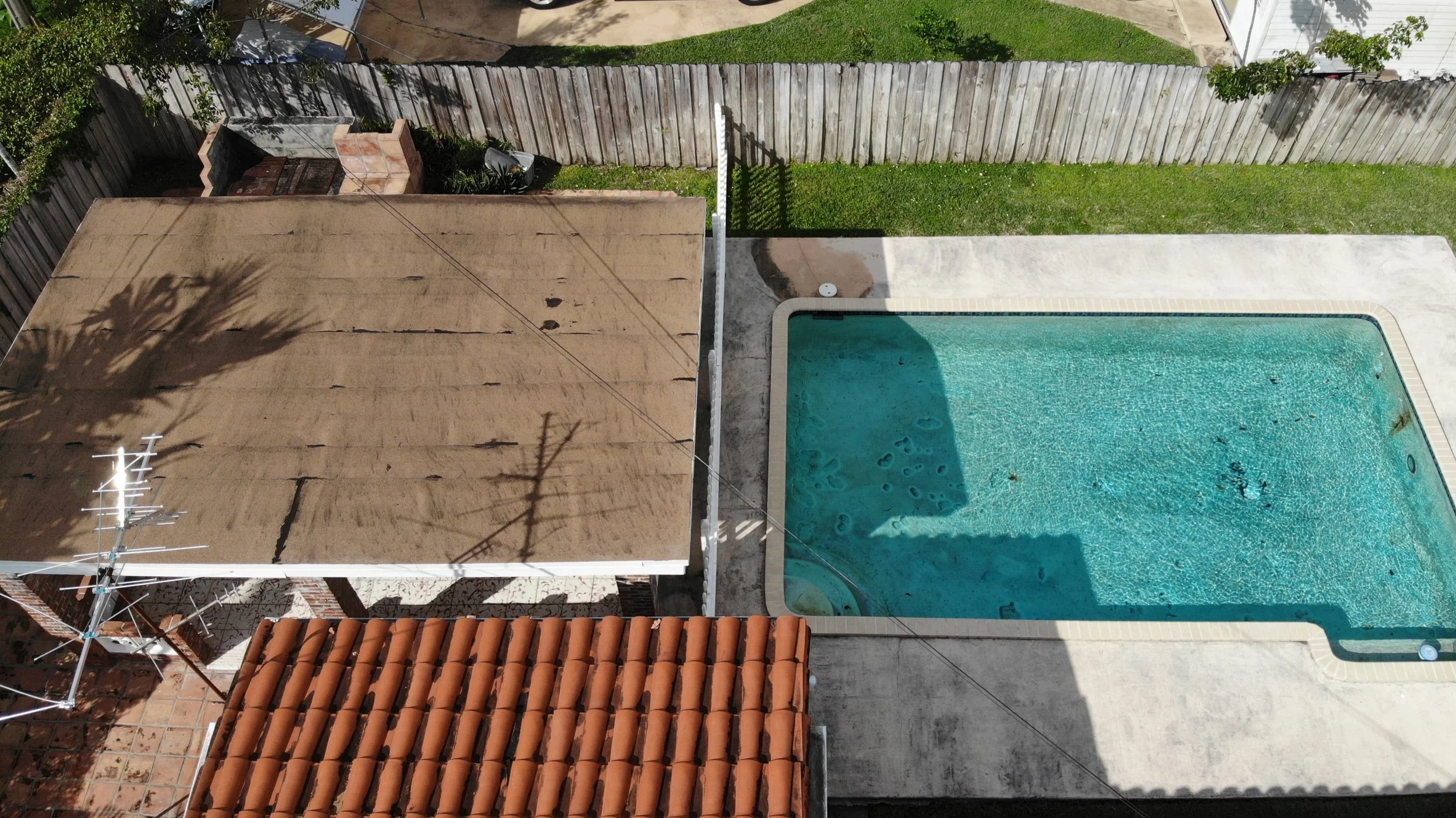 A backyard with a swimming pool, a concrete patio, a fenced yard, and a small grassy area. There is a shaded section on the pool with some leaves and debris on the water.